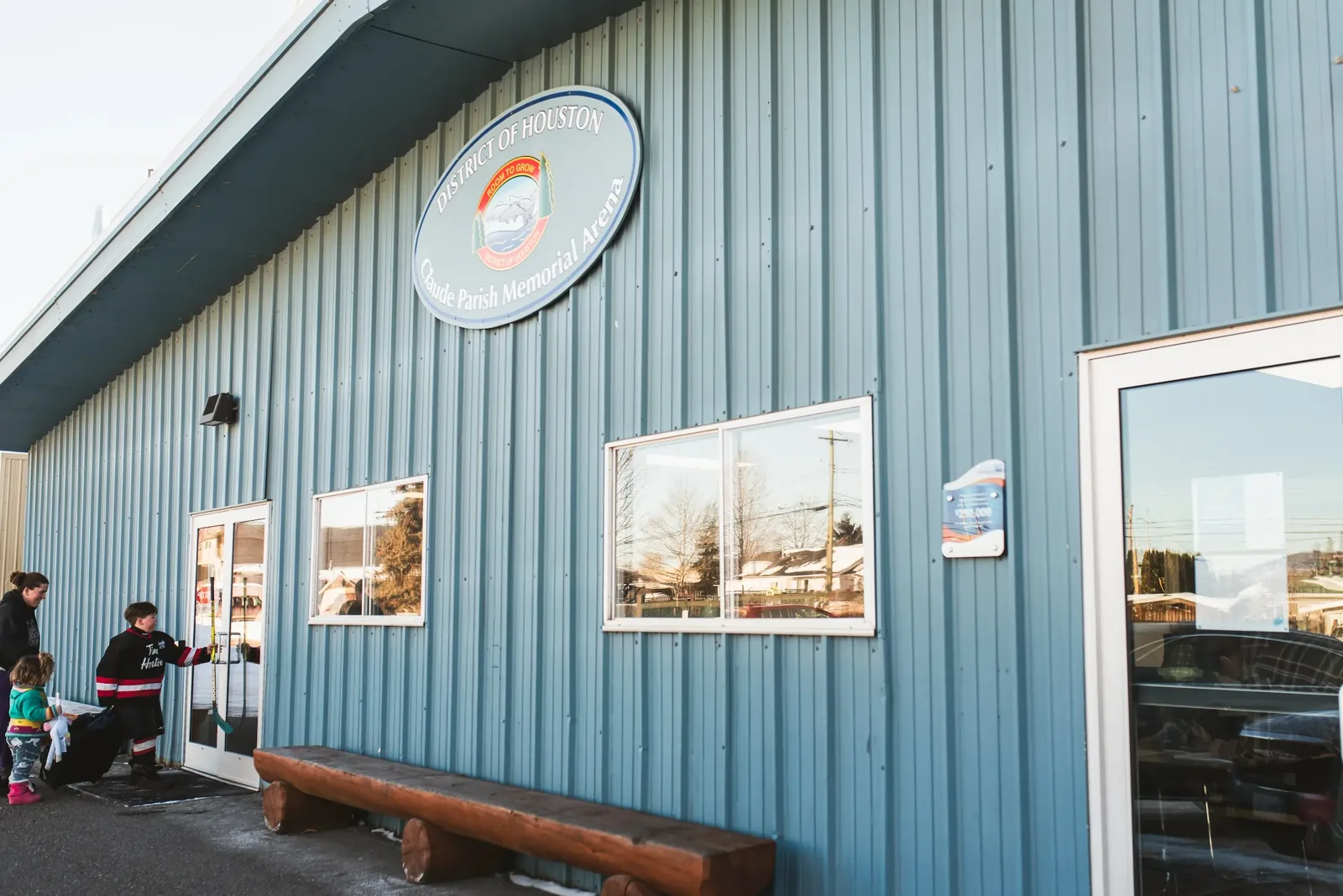 The blue metal front of the Claude Parish Memorial Arena with the District of Houston sign above the doors, a log bench and a few people at the entrance. — Claude Parish Memorial Arena