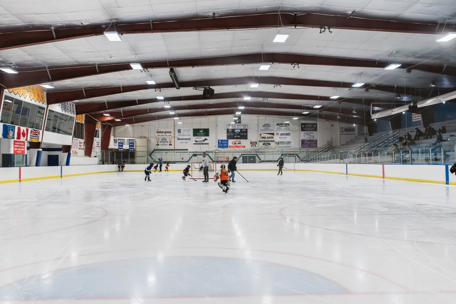 The ice sheet seen from the end of the arena, with a youth practice at the far end, the dasher boards, sponsor signs and a red-and-white steel-truss roof. — Claude Parish Memorial Arena