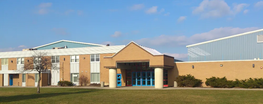 The two-storey brick front of Clearwater Arena with its covered entrance and blue doors, seen from the lawn to the south, with the blue-clad rink halls behind. — Clearwater Arena