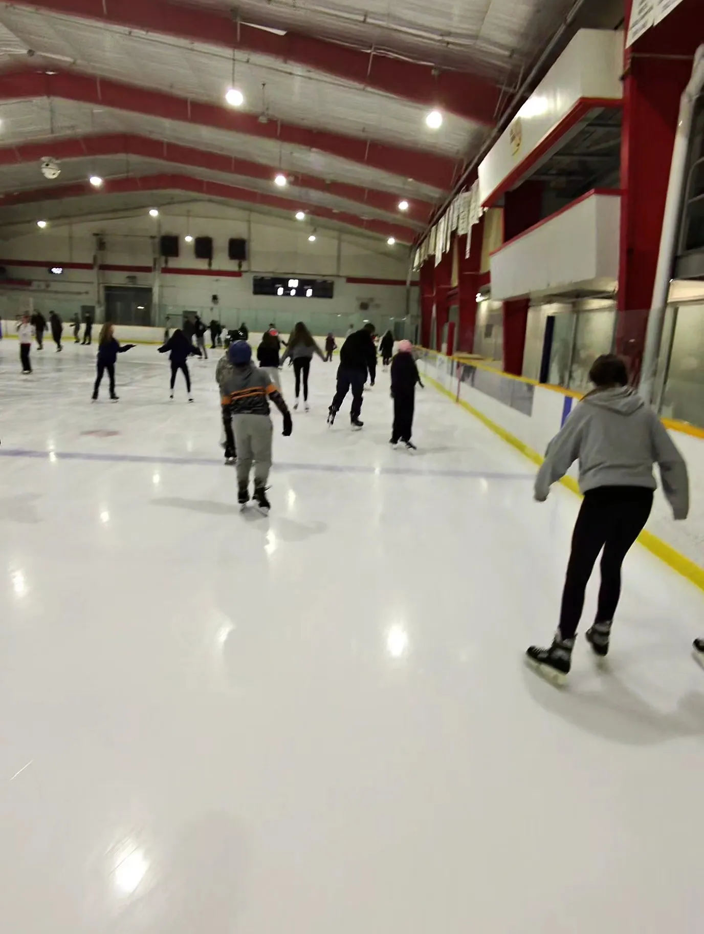 Skaters spread across a wide sheet, seen through a chain-link and glass rail, under a red-trussed steel roof with a scoreboard on the far wall. — Clearwater Arena
