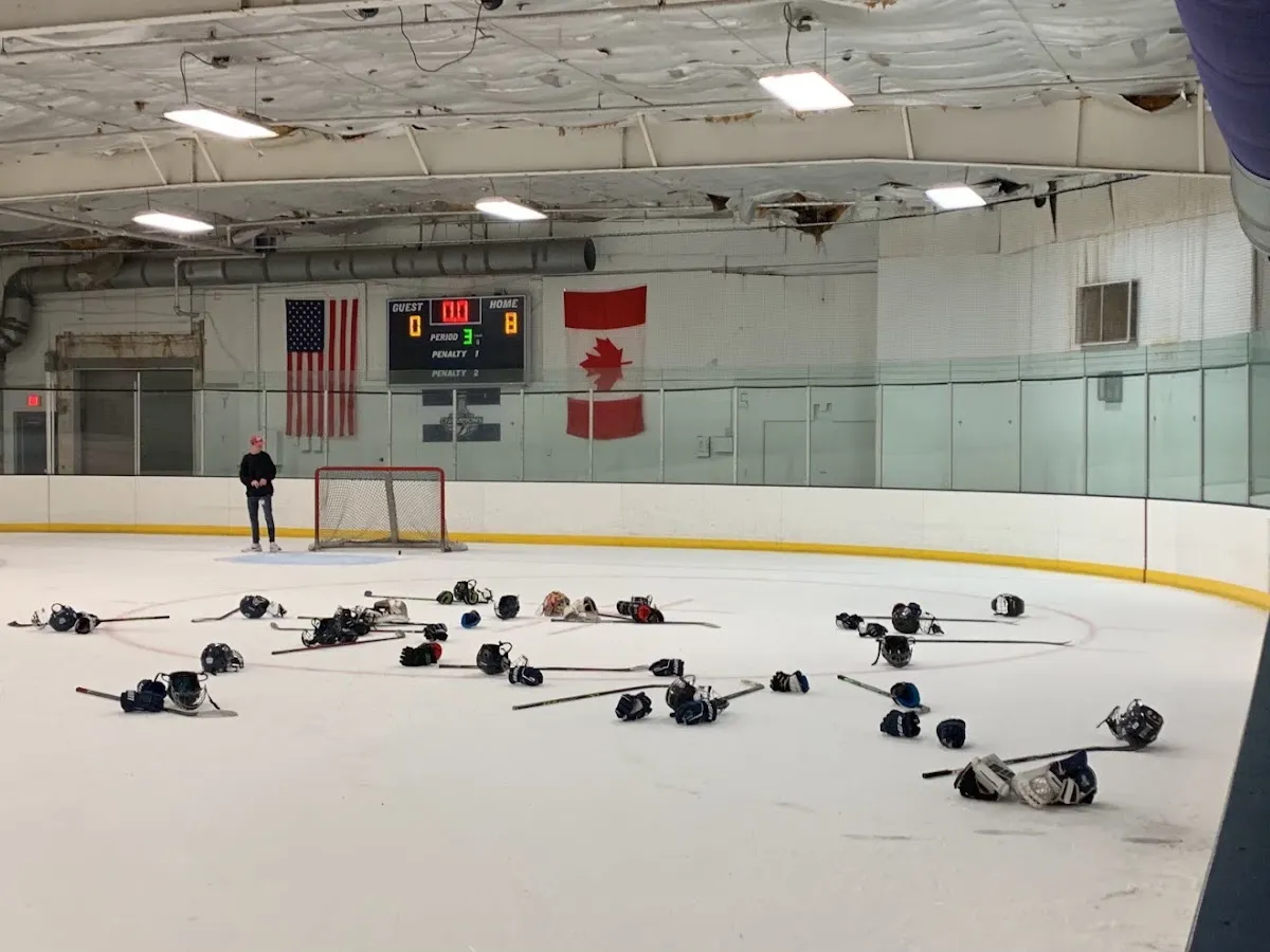 Ice sheet inside the arena with an American and Canadian flag and a scoreboard on the far wall, hockey gear scattered on the ice after a session and one skater in the distance; ceiling insulation above the boards is visibly torn/damaged. — Clearwater Ice Arena