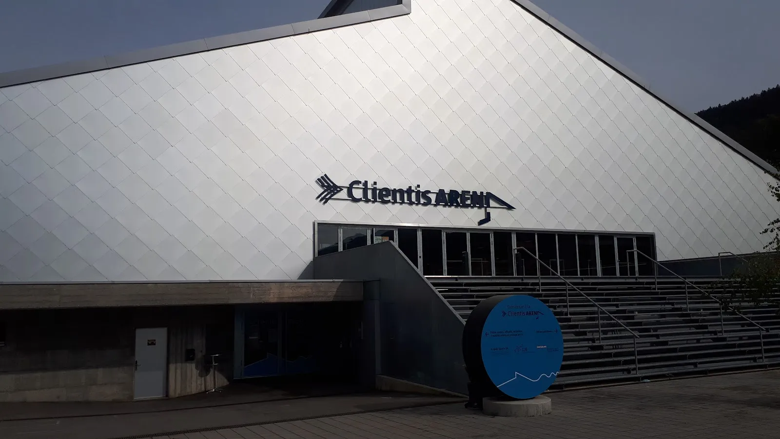 The pale cladding of the Clientis Arena with its name sign above the entrance and a blue drum at the foot of the stairs. — Clientis Arena