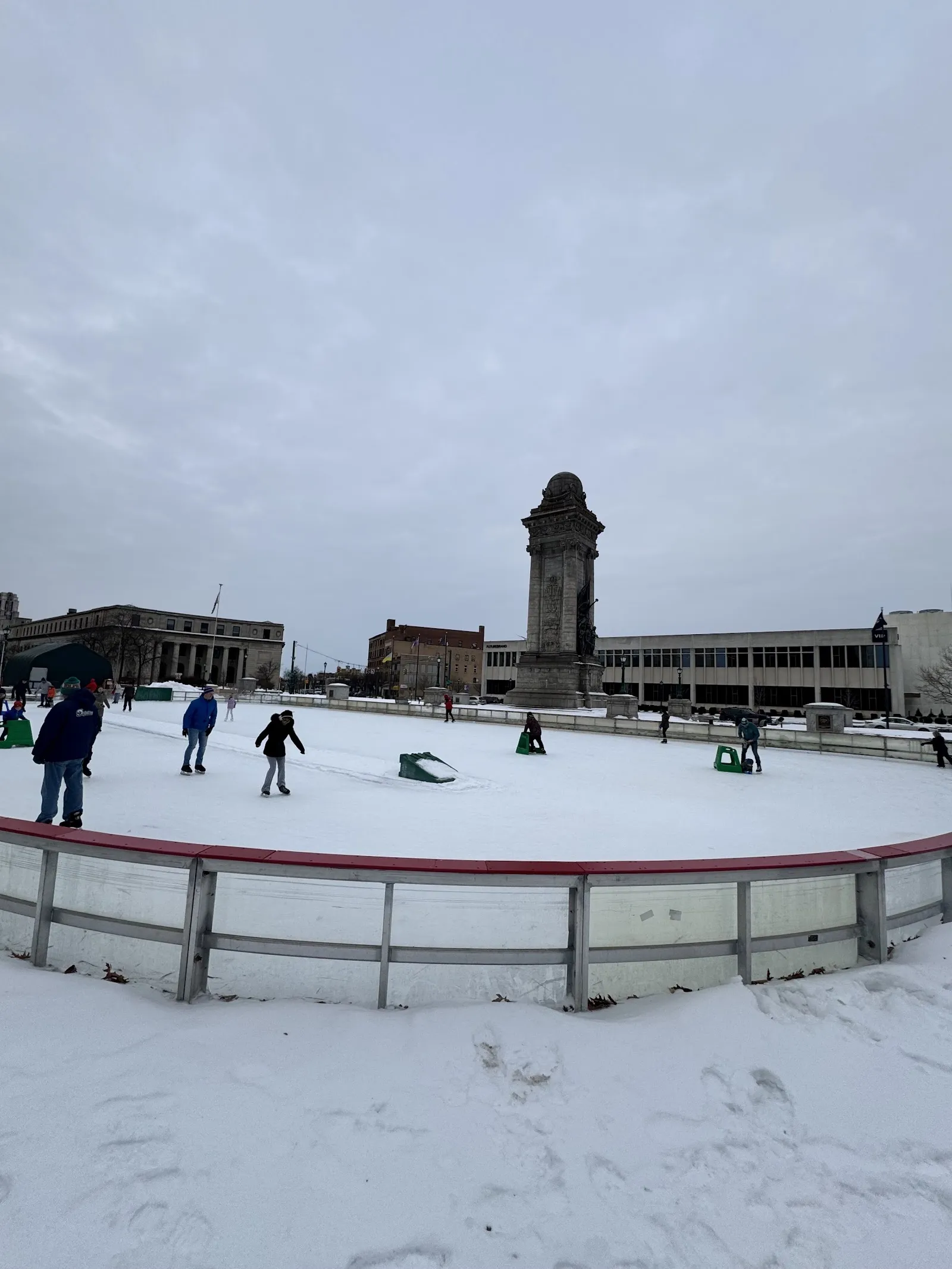 La pista al aire libre de Clinton Square bajo la nieve con unos cuantos patinadores, una alta torre con reloj y los edificios de la plaza del centro de Syracuse detrás. — Clinton Square Ice Rink
