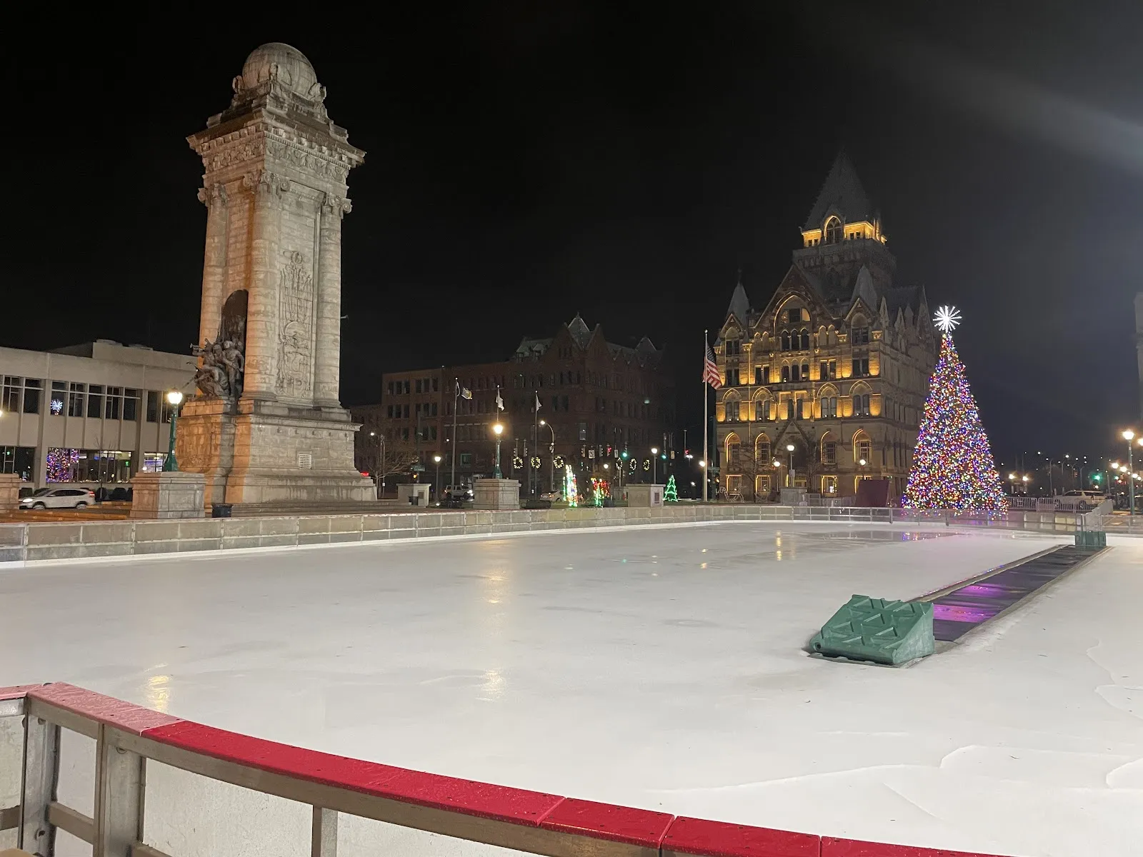 La pista exterior vacía de Clinton Square de noche, con sus vallas de borde rojo, el Soldiers and Sailors Monument y el árbol de Navidad iluminado y el edificio Syracuse Savings Bank al fondo — Clinton Square Ice Rink