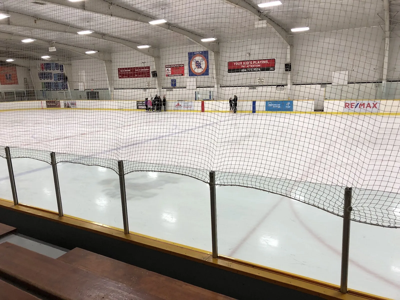 The Cloverdale Arena ice from the wooden bleachers, through the netting toward the far boards and lacrosse banners — Cloverdale Arena