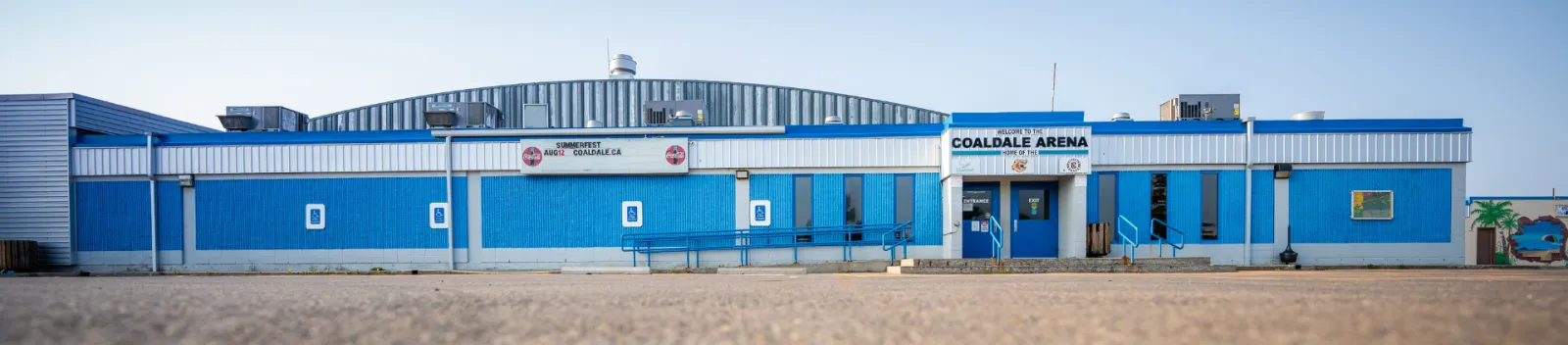 The long, low blue-and-white Coaldale Arena building with its name over the entrance, seen across a gravel lot. — Coaldale Arena