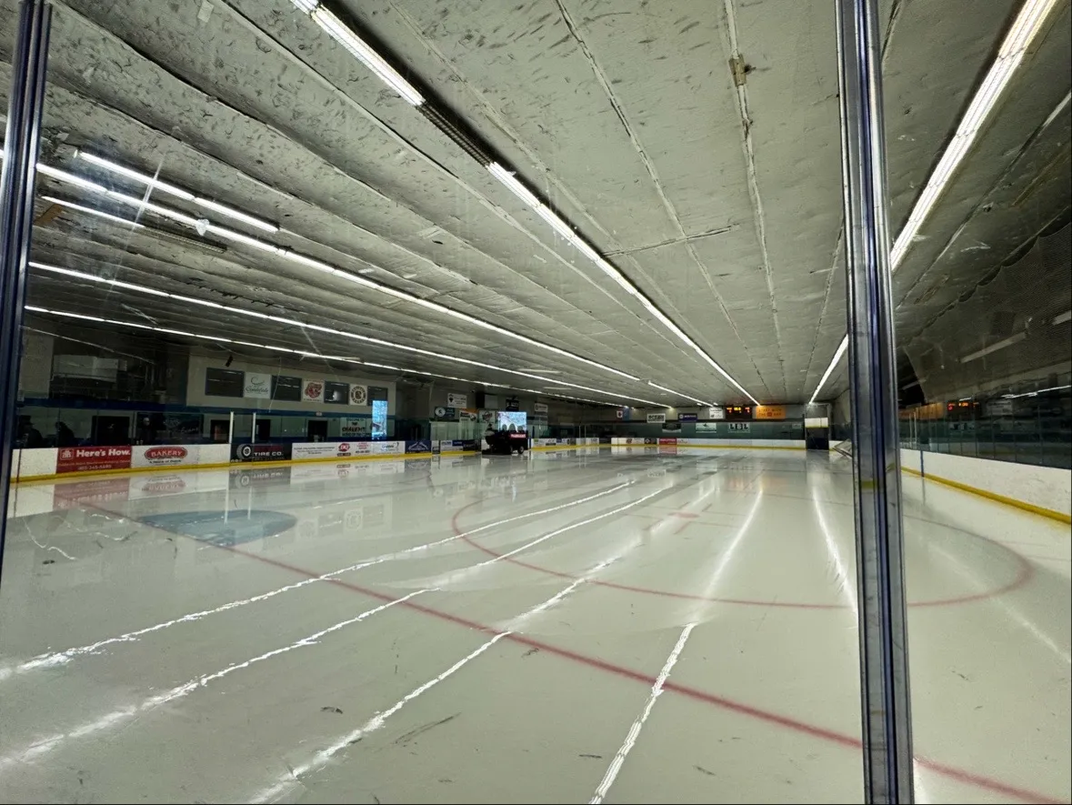 The empty ice at the Coaldale Arena, seen through the glass along the boards toward the far end and scoreboard — Coaldale Arena