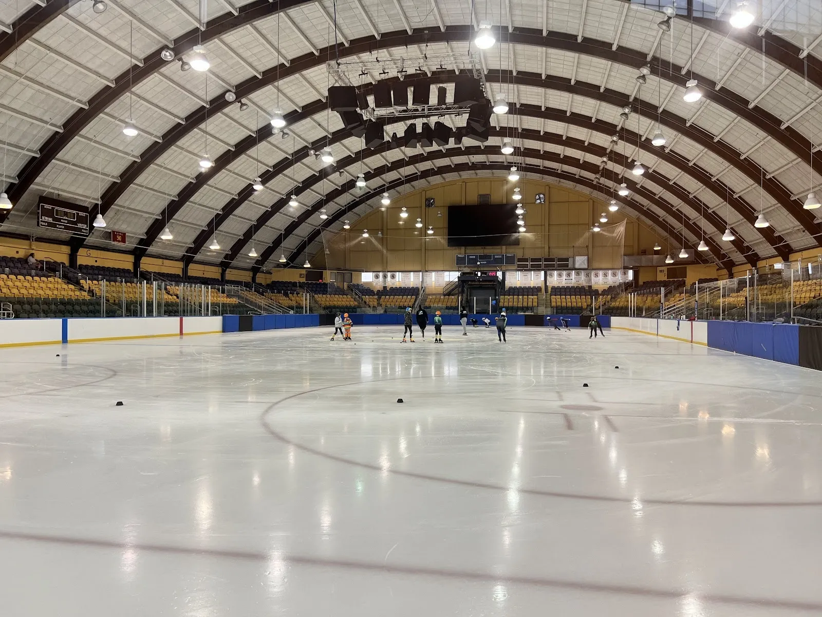 El hielo vacío bajo un amplio techo arqueado con costillas color madera, con un círculo central y vallas azules, y gradas a lo largo del lado opuesto. — Essex County Richard J. Codey Arena