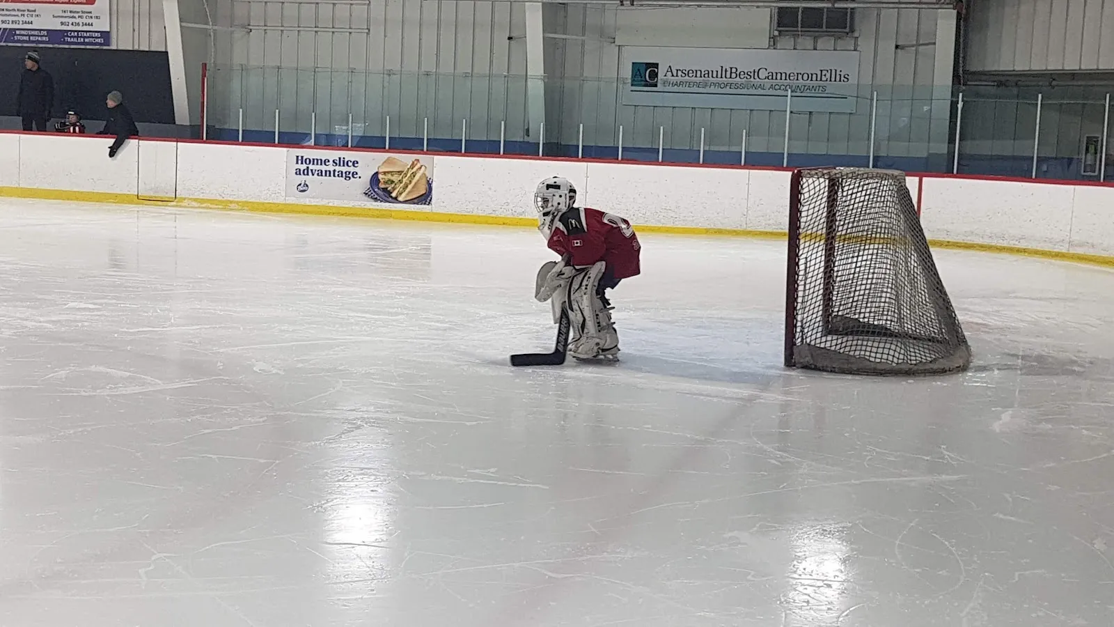 A goalie crouched beside the net on a clean white sheet of ice at Cody Banks Arena, with sponsor boards and the pale blue upper wall behind. — Cody Banks Arena