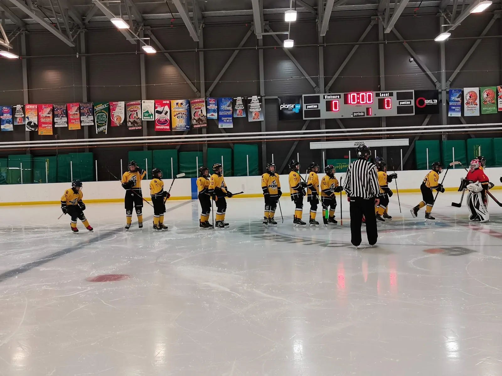 Une équipe de jeunes alignée sur la glace avec un arbitre au Colisée Béton-Provincial, avec le tableau indicateur et la rangée de bannières de commanditaires au-dessus des bandes. — Colisée Béton-Provincial