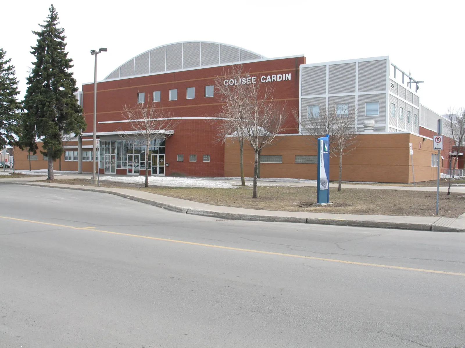 The Colisée Cardin from the street: a red-brick front with 'COLISÉE CARDIN' lettering, the curved roof of the arena rising behind it and a blue Sorel-Tracy sign post on the lawn. — Colisée Cardin