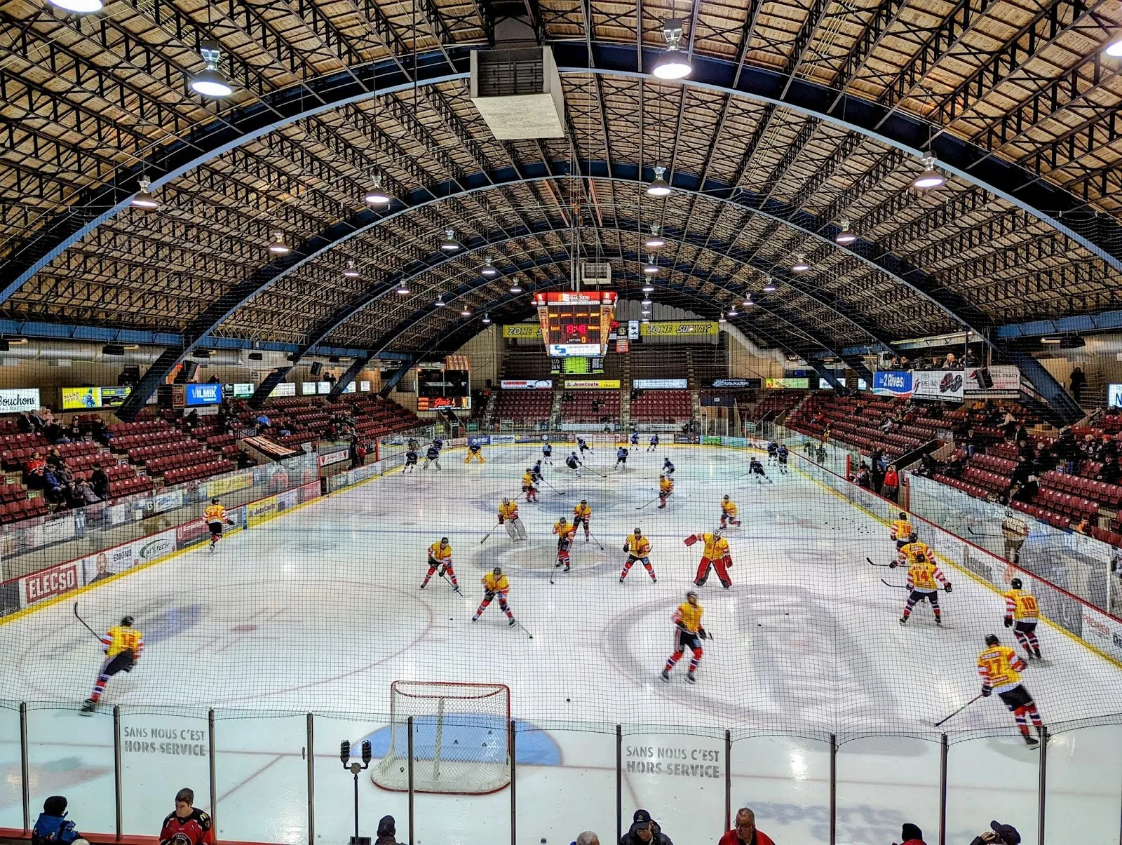 The arched grandstand arena seen from the end, players in yellow and red spread across the sheet under a wide timber-ribbed roof. — Colisée Cardin