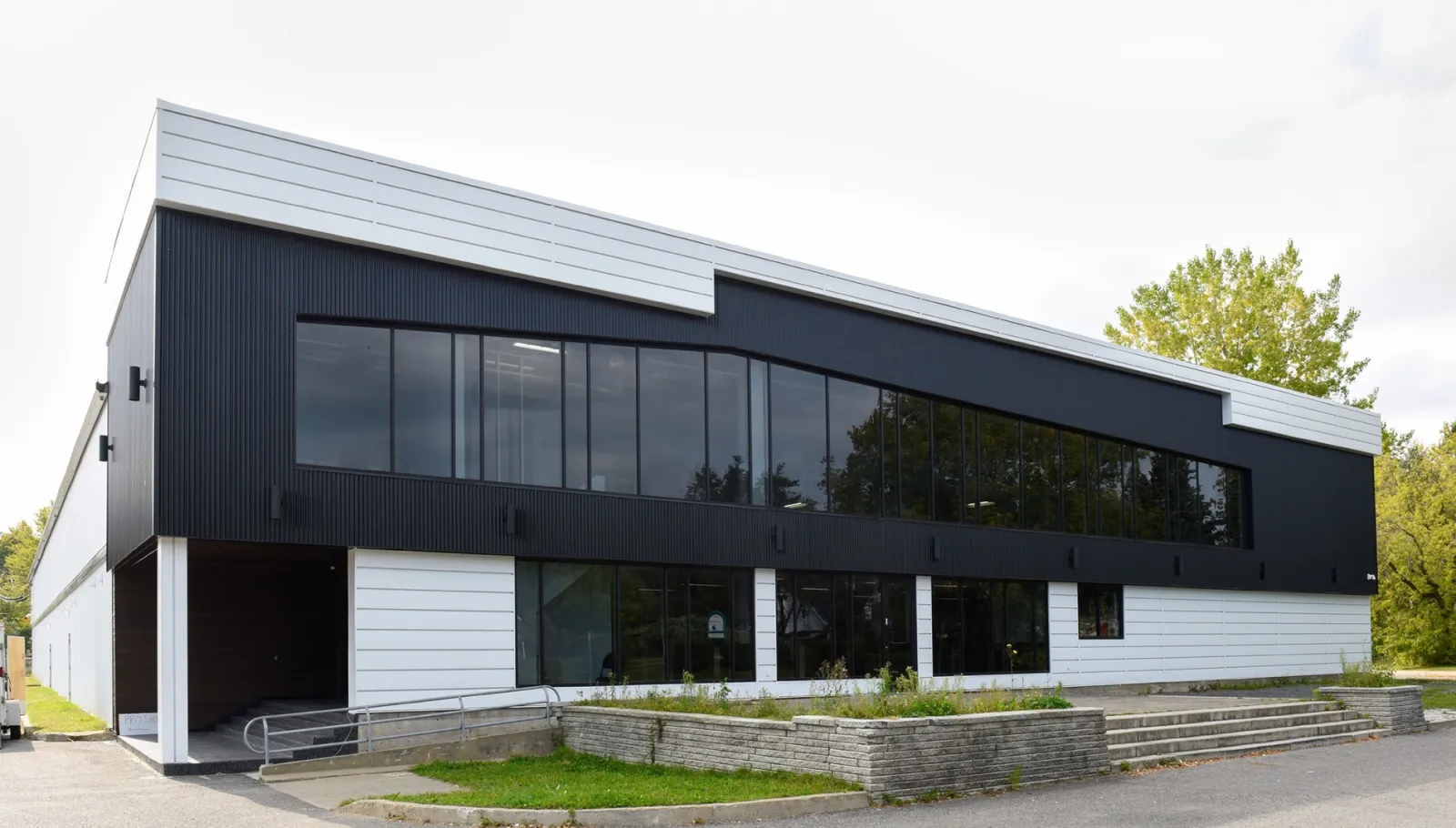 La façade de l’aréna : un étage supérieur revêtu de noir, percé d’une longue bande de fenêtres, au-dessus d’un rez-de-chaussée blanc, avec des marches et une rampe menant à l’entrée. — Colisée de Loretteville