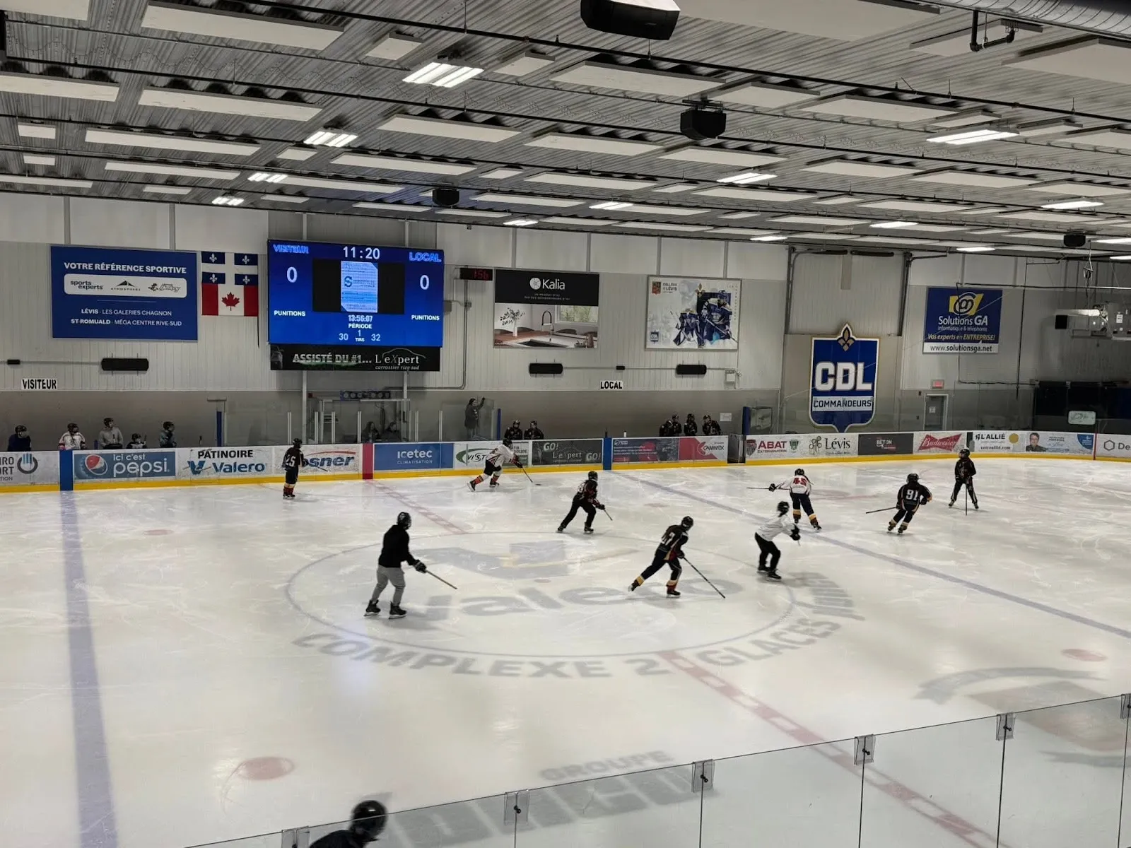 Un entraînement sur une grande surface de glace sous un tableau indicateur bleu et un mur de bannières, des joueurs répartis sur la glace autour d’un logo central. — Colisée de Loretteville