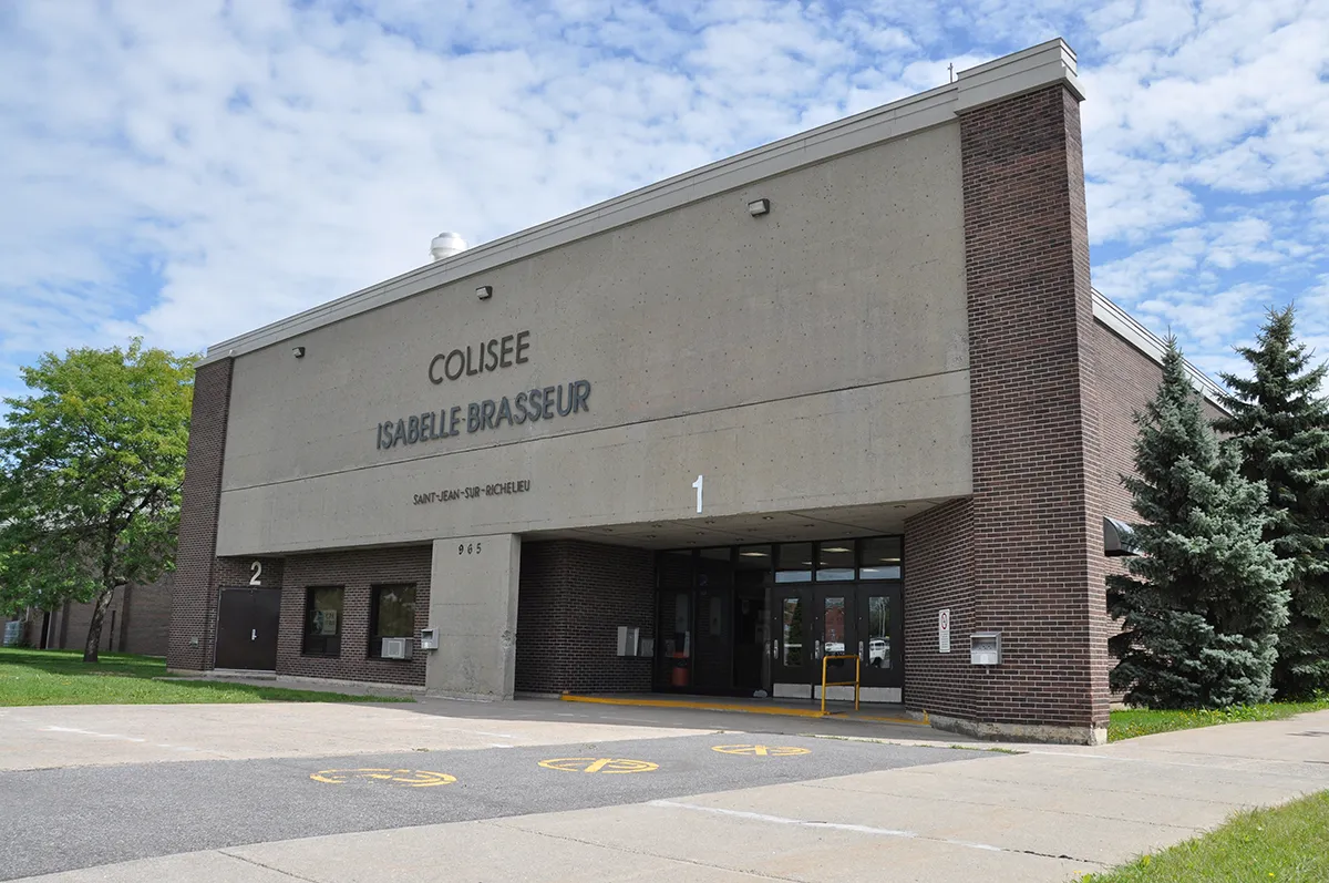 La fachada de bloques grises de la arena con las letras de Colisée Isabelle-Brasseur, con muros laterales de ladrillo, dos entradas numeradas y coníferas al lado. — Colisée Isabelle-Brasseur