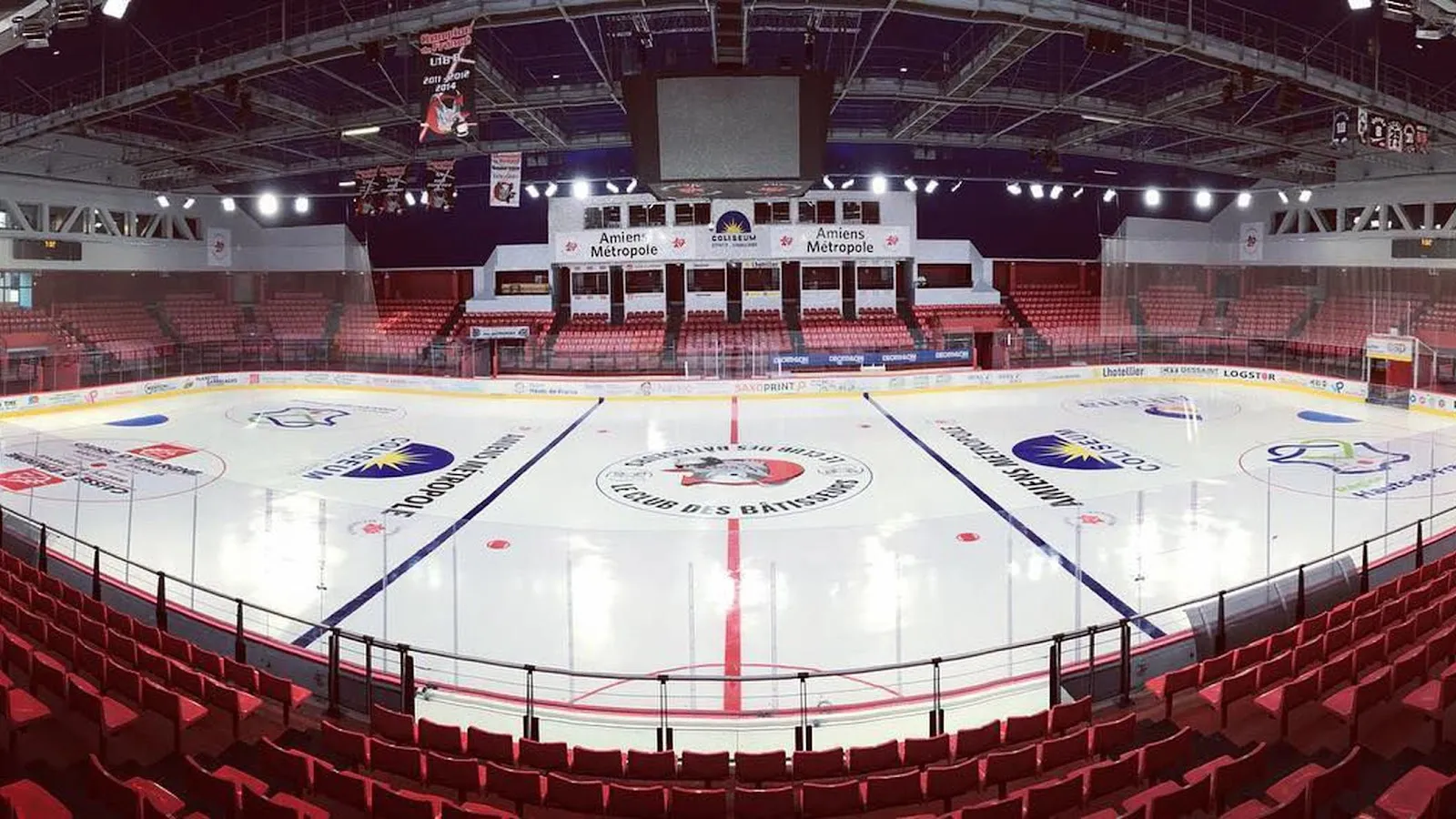 Vue panoramique de la glace vide de l’arène du Coliseum depuis les gradins, avec des sièges rouges tout autour, les bannières d’Amiens Métropole et le tableau d’affichage central. — Coliseum, patinoire