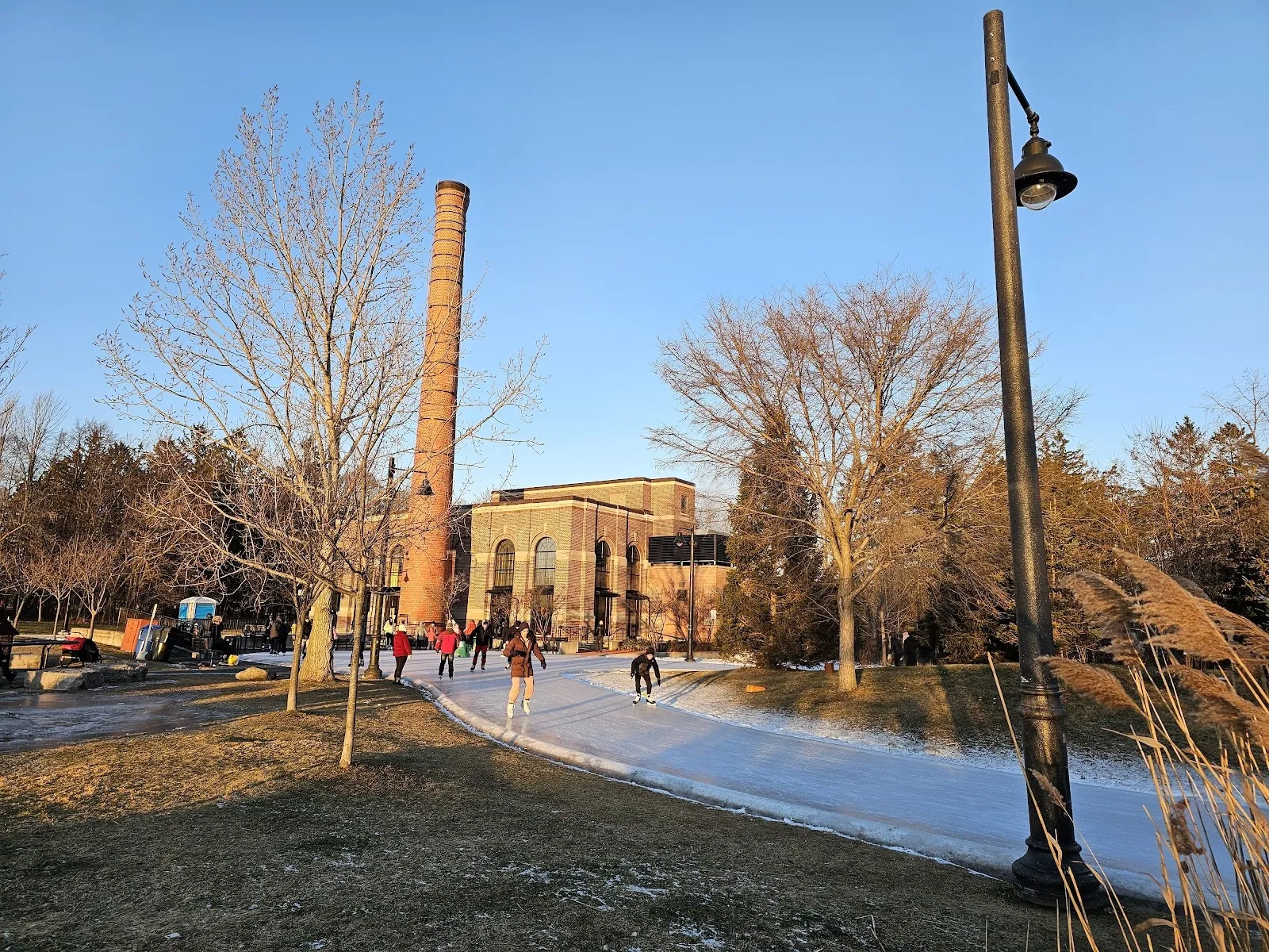 The brick building with the tall chimney beside the Colonel Samuel Smith skate trail, with a curve of ice, a lamp post and a few skaters in front under a clear evening sky. — Colonel Samuel Smith Park Skate Trail