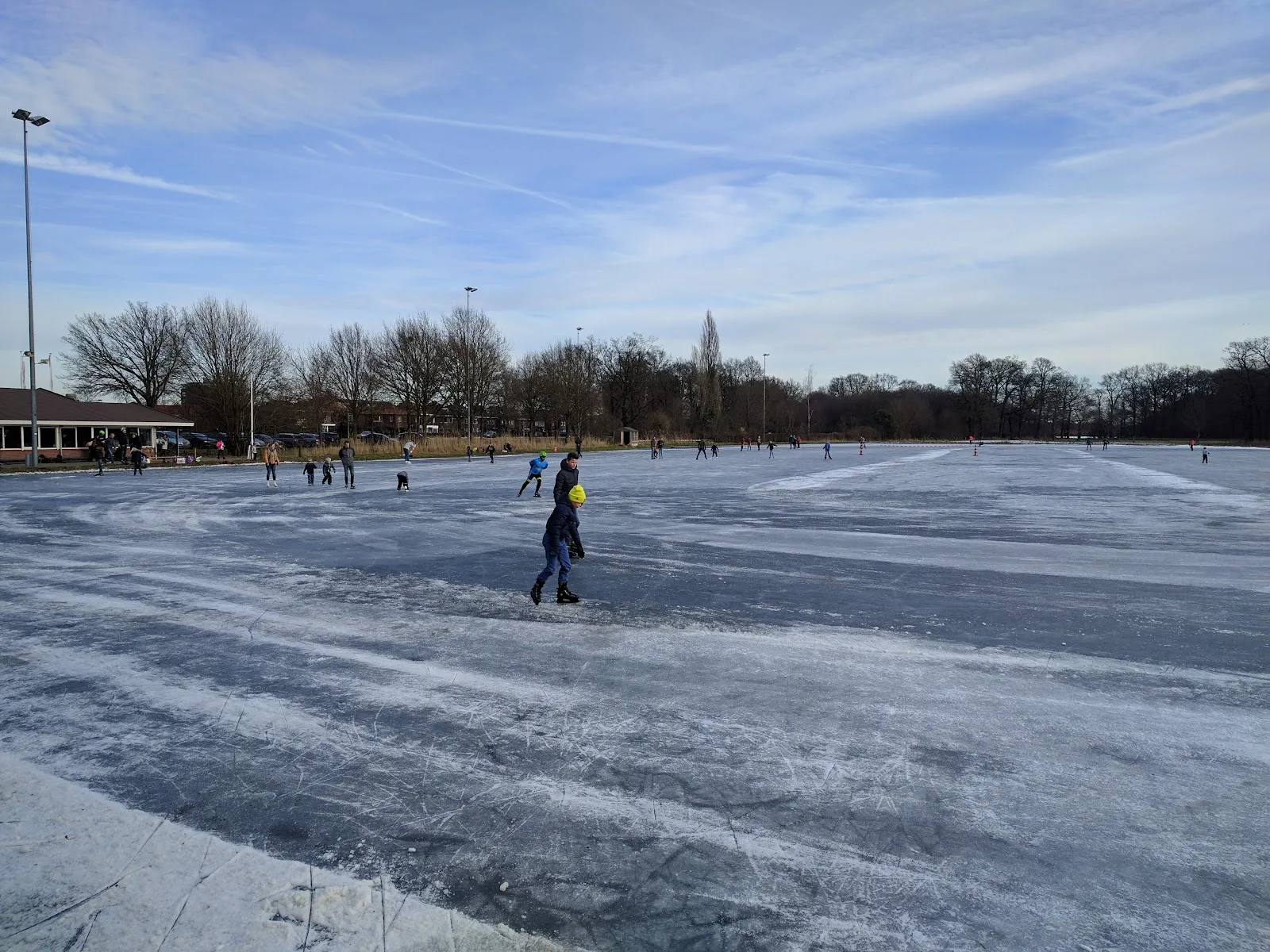 Skaters spread across the wide open natural-ice track at Combibaan Hengelo on a sunny winter day, with bare trees and the clubhouse behind. — Combibaan Hengelo