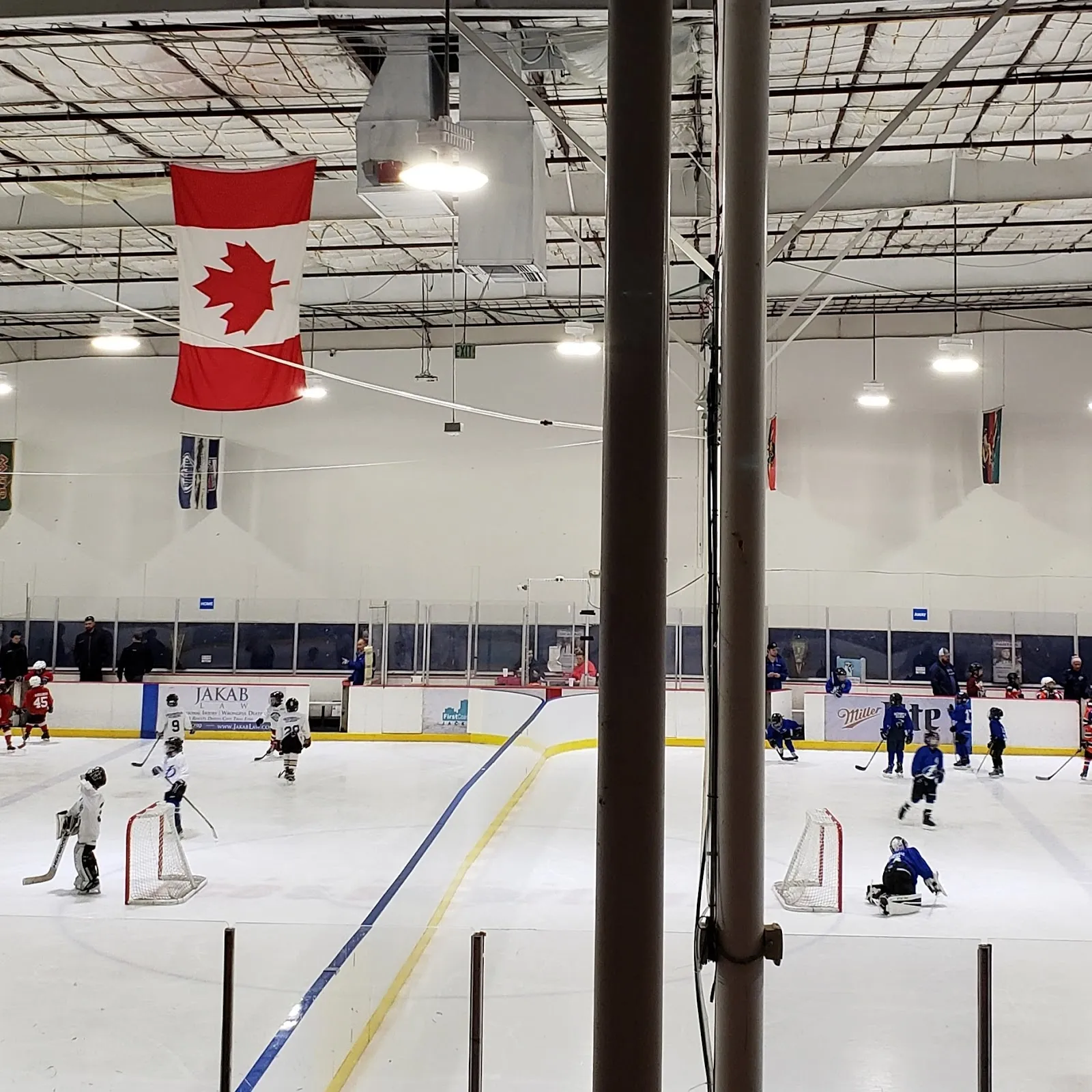 Una sesión pública vista a través de un poste y vidrio desde las gradas, con patinadores y jóvenes jugadores repartidos por el hielo y una bandera de Canadá colgando del techo. — Community First Igloo
