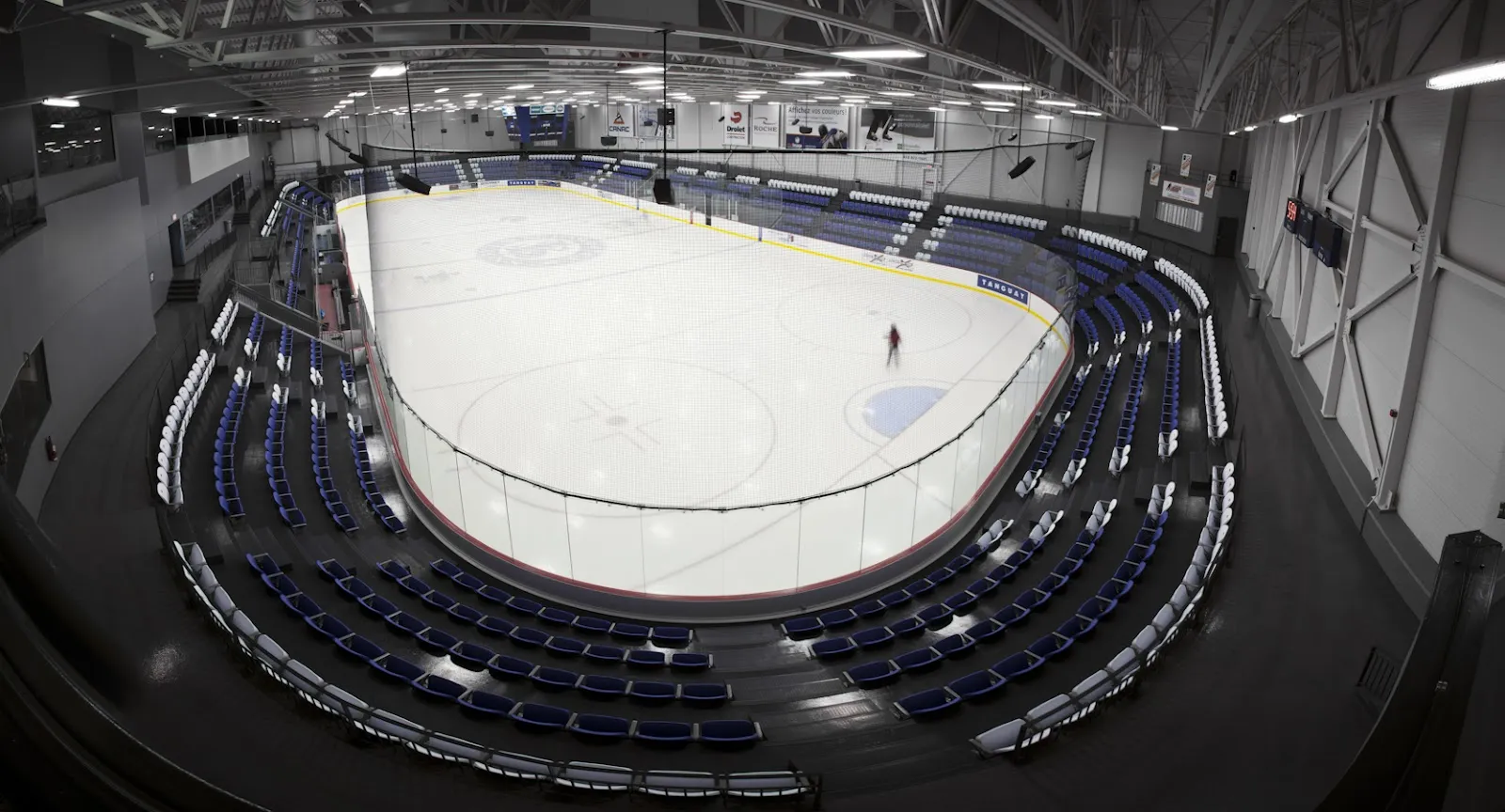 Una vista amplia y elevada de la pista principal: toda la pista rodeada de hileras de asientos azules y un pasillo arriba, con un patinador en el hielo. — Complexe sportif Bonair