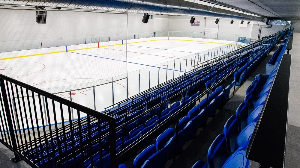 La surface de glace vide du Complexe sportif de Paspébiac vue des gradins, avec des rangées de sièges bleus le long des bandes et les murs et le plafond blancs du nouvel aréna. — Complexe sportif de Paspébiac
