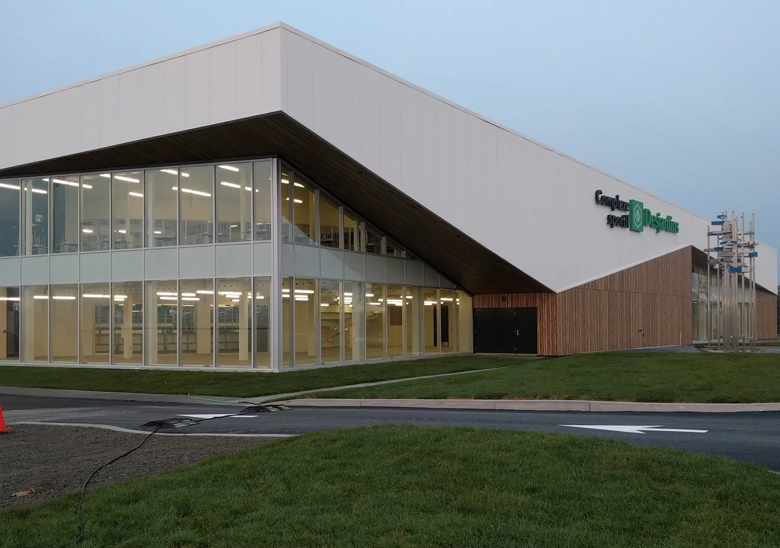 Le nouveau bâtiment de verre et de panneaux blancs du Complexe sportif Desjardins d’Amos au crépuscule, avec son nom sur le mur du bout et une base revêtue de bois. — Complexe sportif Desjardins d'Amos