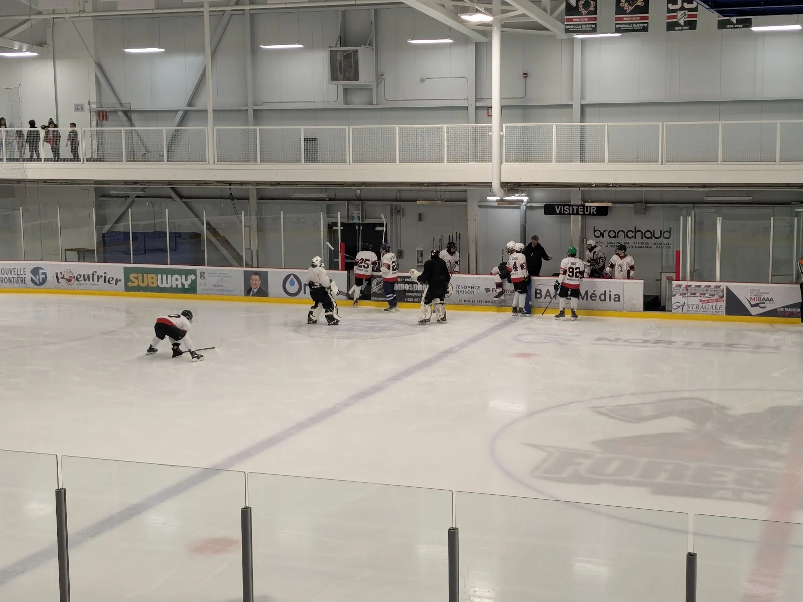 Des joueurs sur la glace du Complexe sportif Desjardins d’Amos, avec les panneaux de commanditaires le long de la baie vitrée et une passerelle en mezzanine au-dessus des bandes du fond. — Complexe sportif Desjardins d'Amos
