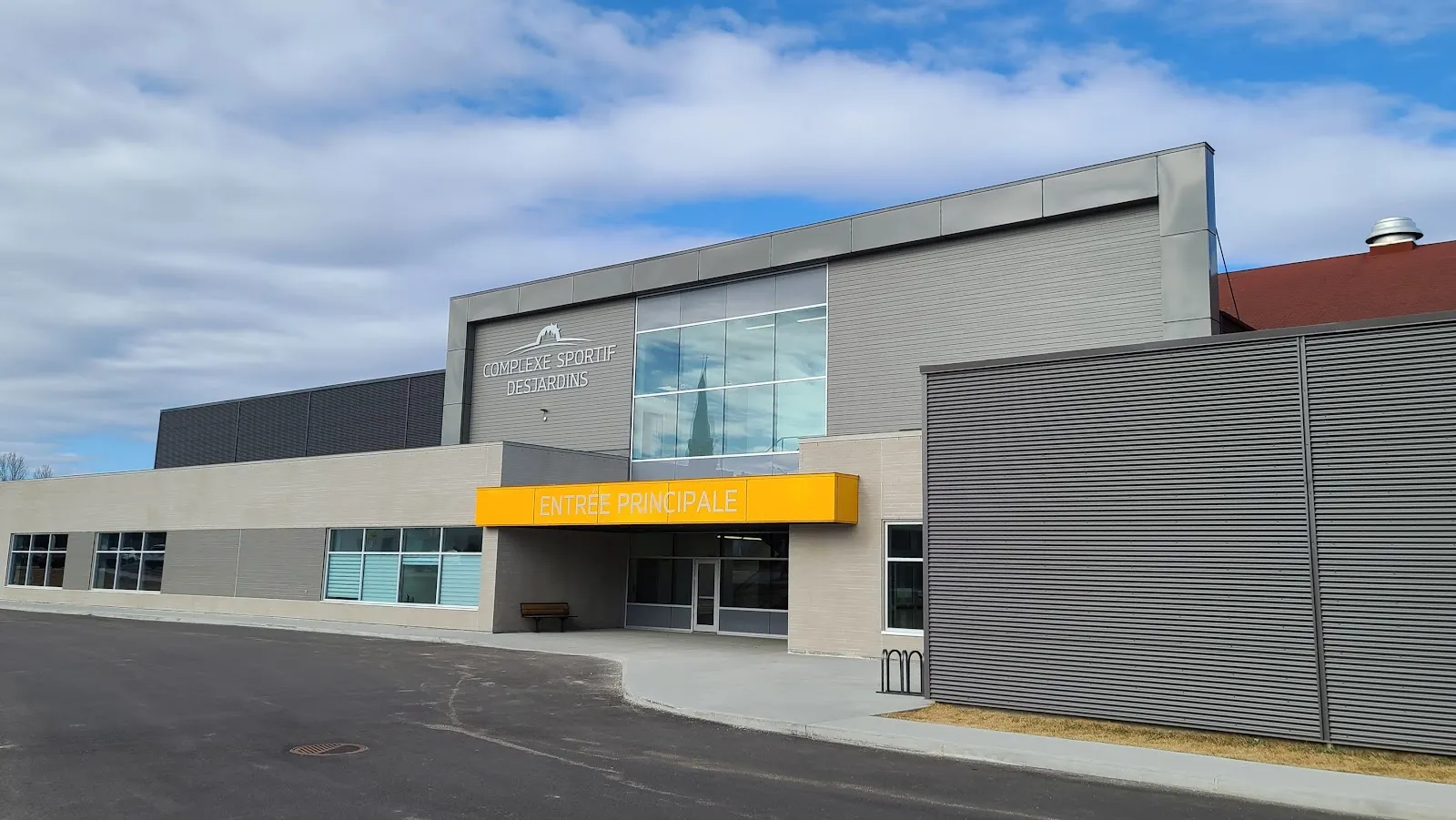Le Complexe sportif Desjardins : un volume gris avec une marquise jaune au-dessus de l’entrée principale et des murs de métal nervuré. — Complexe sportif Desjardins de Dolbeau-Mistassini