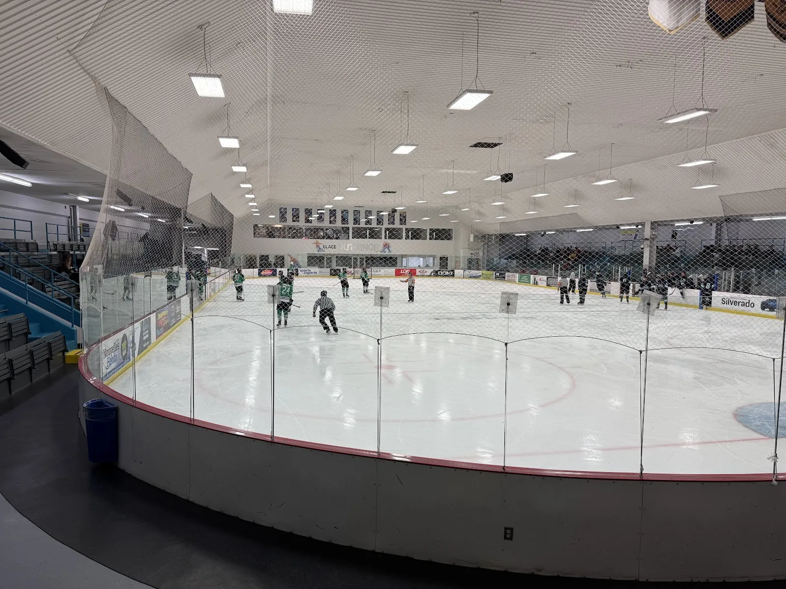 Une vue large de la glace par-dessus une rampe courbe, quelques joueurs répartis sur la glace sous un haut plafond, avec un mur de verre éclairé à gauche. — Complexe sportif Desjardins de Dolbeau-Mistassini