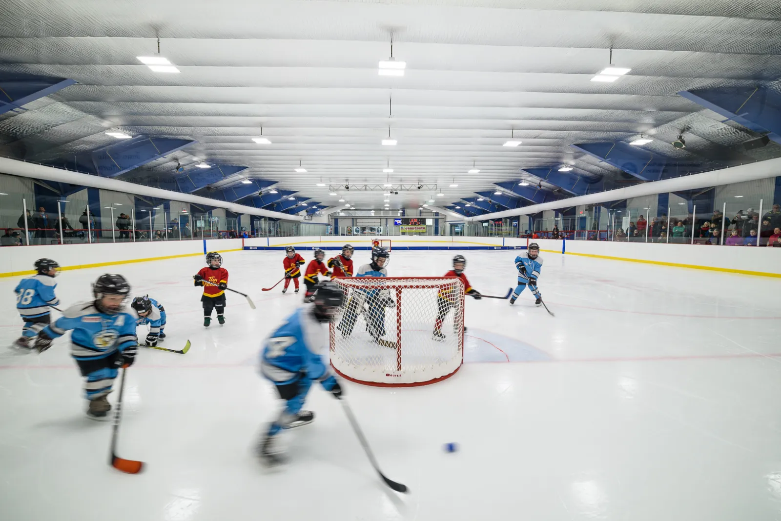 De jeunes joueurs sur la glace de l’aréna de Grande-Rivière pendant une partie de hockey mineur, avec des spectateurs derrière la baie vitrée des deux côtés et le plafond bleu et blanc au-dessus. — Aréna de Grande-Rivière