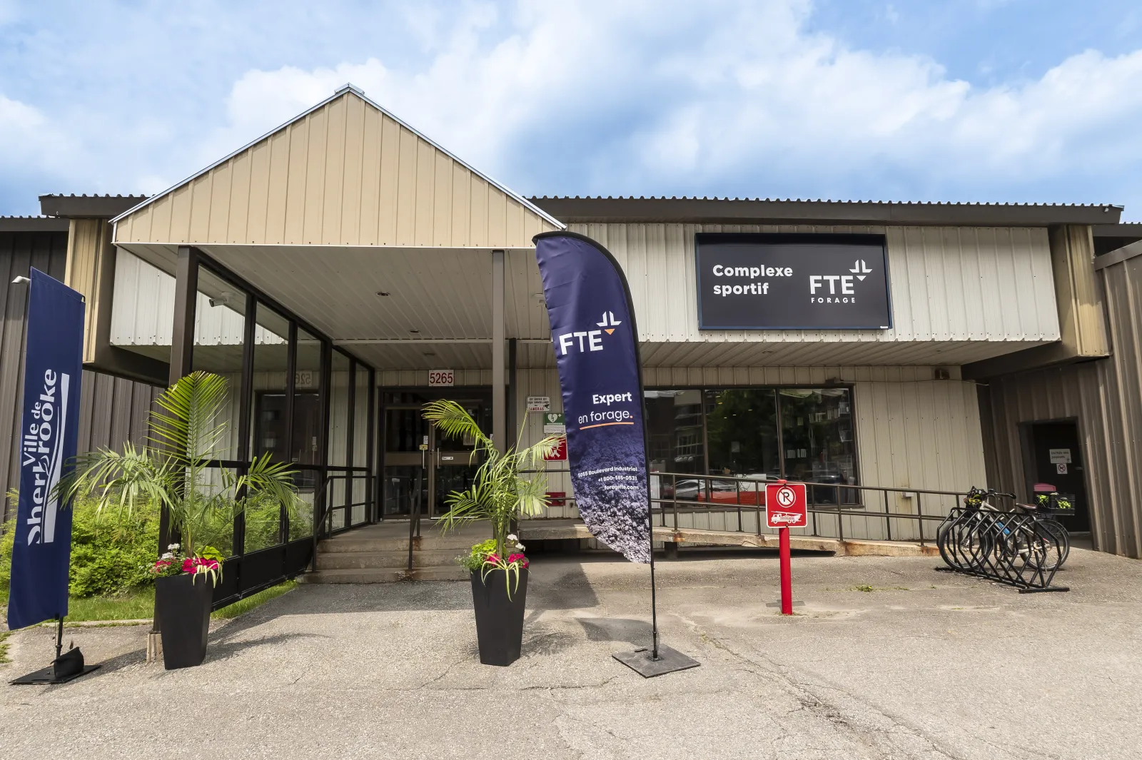 L’entrée vitrée à pignon du bâtiment revêtu de métal crème, portant l’enseigne « Complexe sportif FTE Forage », avec des bannières et des jardinières devant. — Complexe sportif Forage FTE