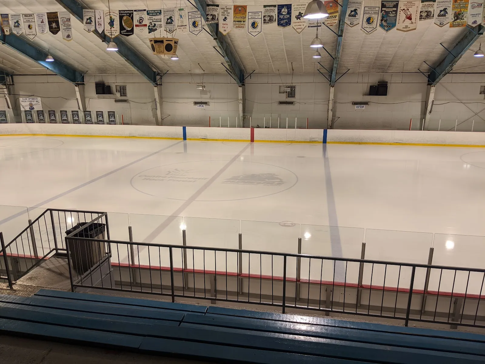 La patinoire vide vue des gradins au centre de la glace, avec des rangées de fanions d’équipes suspendus sous les fermes d’acier du toit. — Complexe sportif Forage FTE
