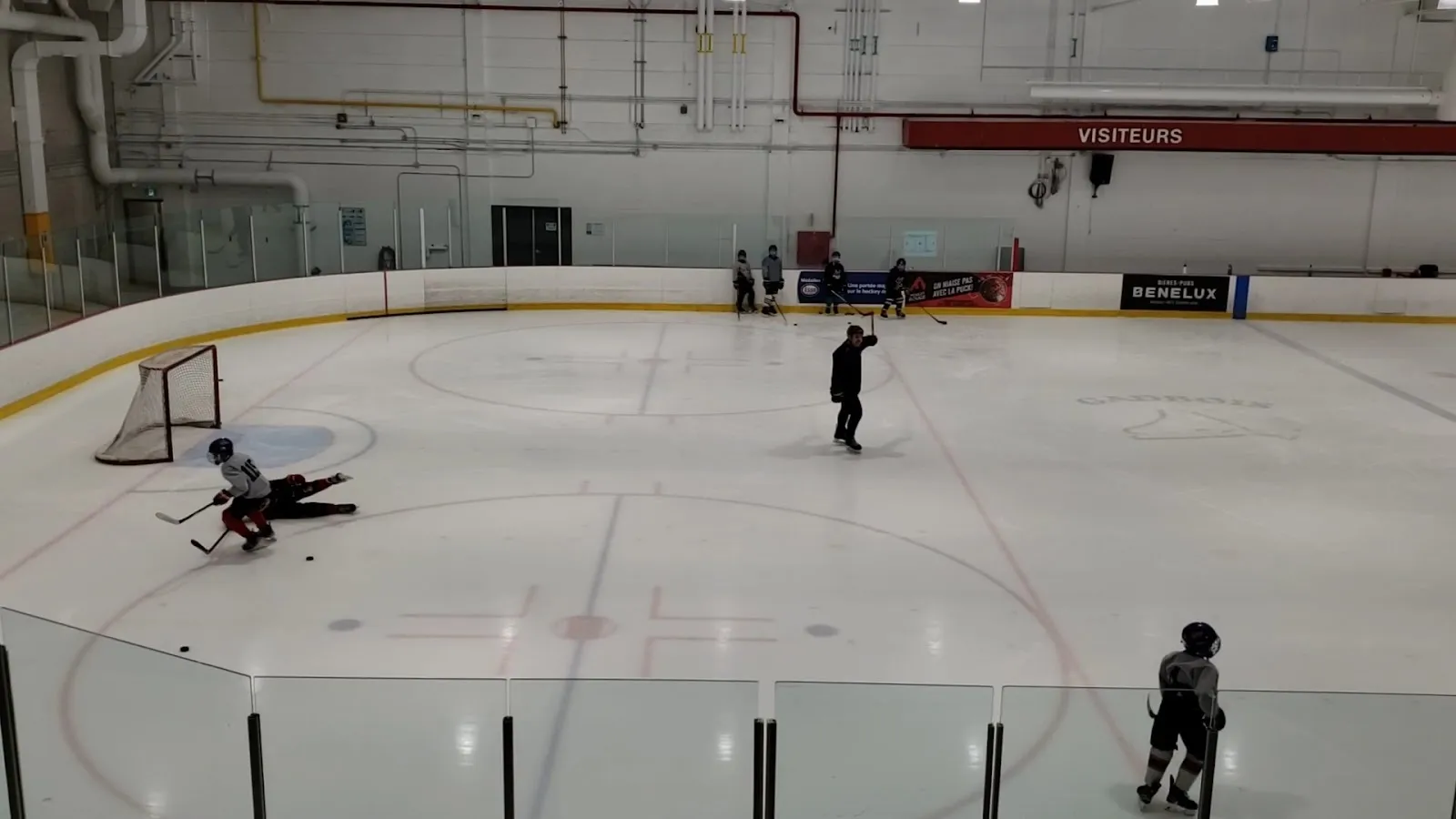 Un entraînement à une extrémité de la glace vu à travers la baie vitrée, des joueurs répartis près d’un filet sous un mur de bannières rouges et blanches. — Complexe sportif Gadbois