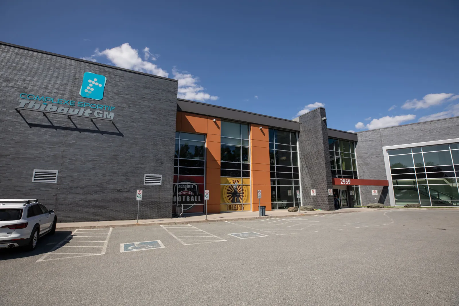 La façade gris foncé et vitrée du complexe, portant l’enseigne « Complexe sportif Thibault GM », à côté d’un stationnement doté de places accessibles. — Complexe sportif Thibault GM