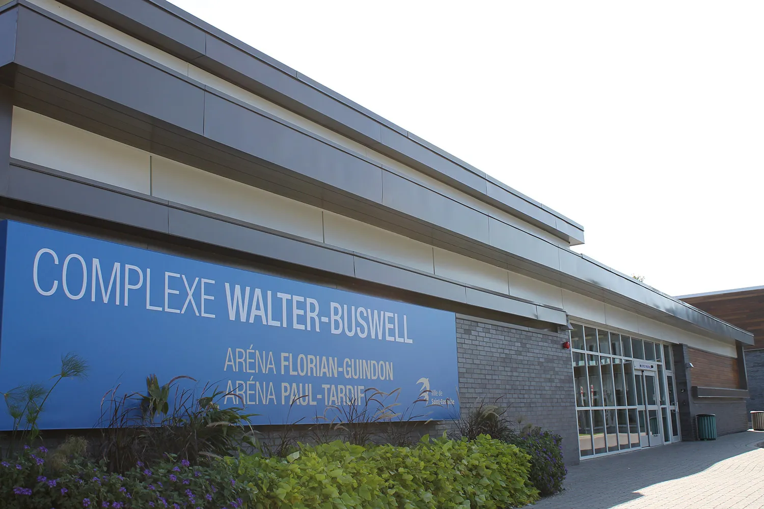 El frente de la entrada del complejo, con un letrero azul que dice Complexe Walter-Buswell, Aréna Florian-Guindon, Aréna Paul-Tardif junto a puertas de vidrio. — Complexe Walter-Buswell
