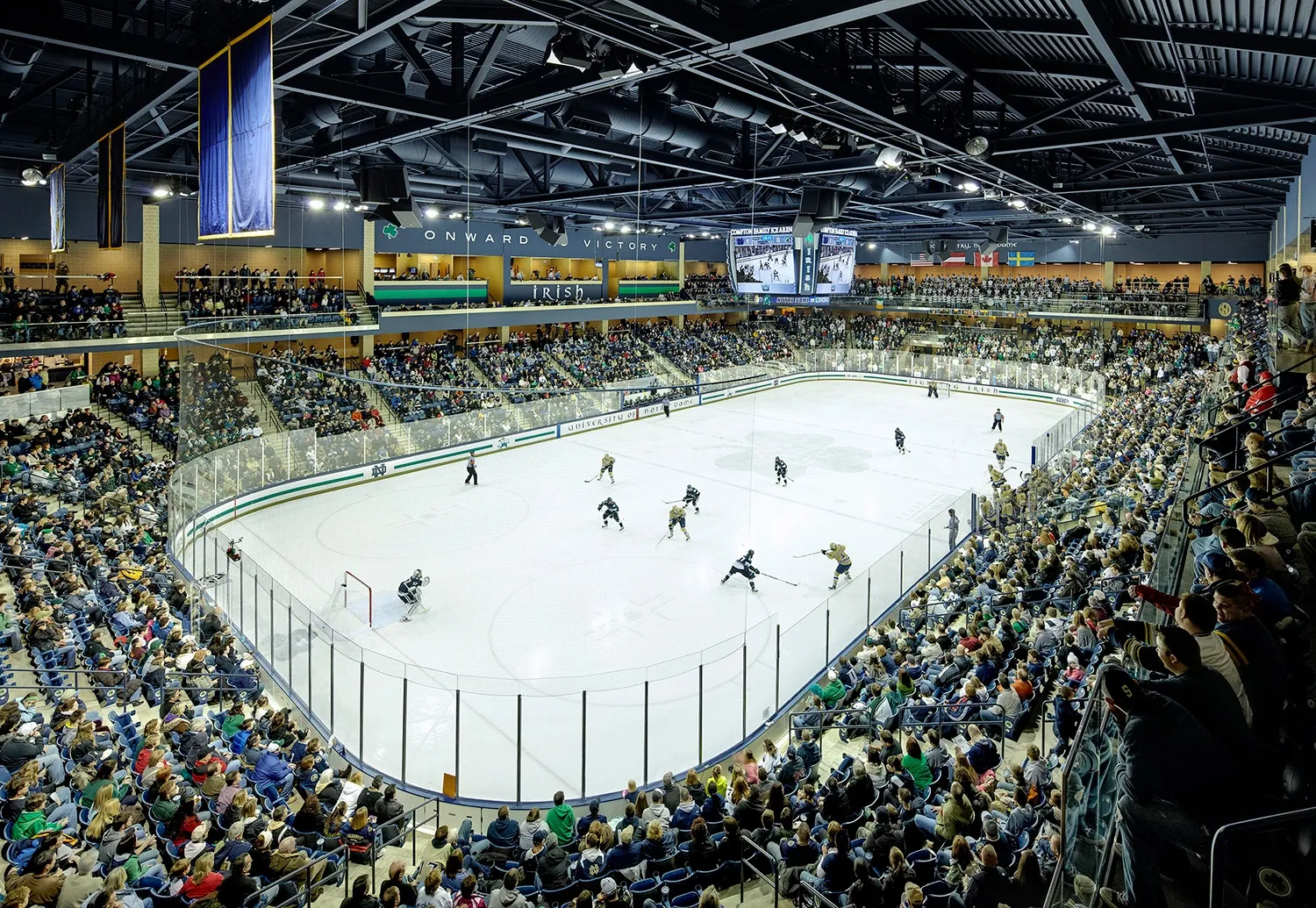 The arena bowl from the upper seats, a game in play on the sheet with dark blue seating ringing the ice and a steel-trussed roof overhead. — Compton Family Ice Arena