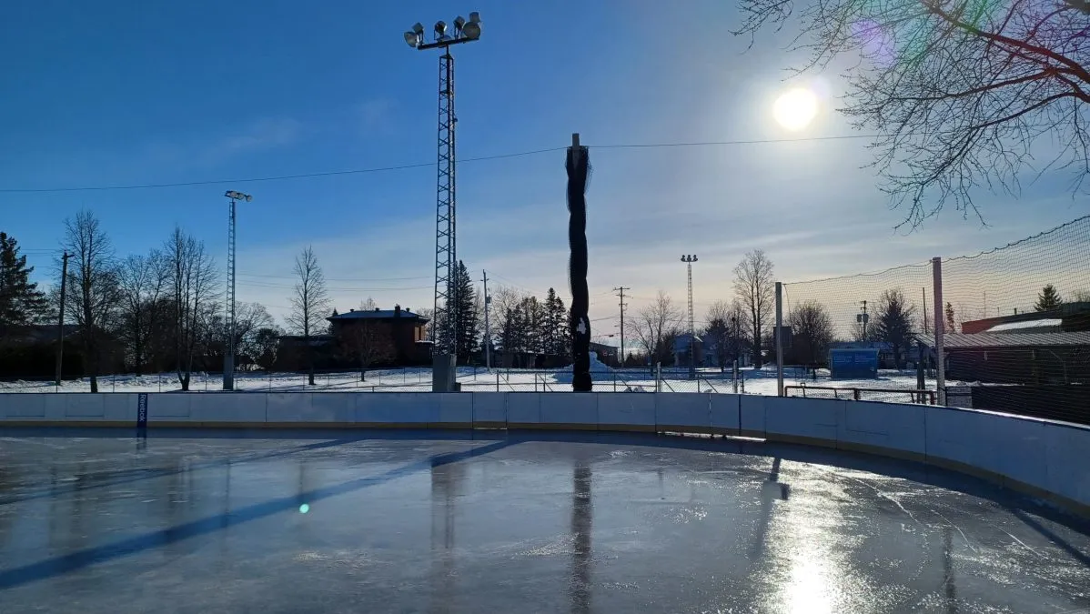 Las vallas blancas curvas y el hielo liso de la pista exterior de Conestogo bajo el sol bajo del invierno, con torres de focos, redes y casas más allá de la cerca. — Conestogo Park Outdoor Rink (Woolwich)