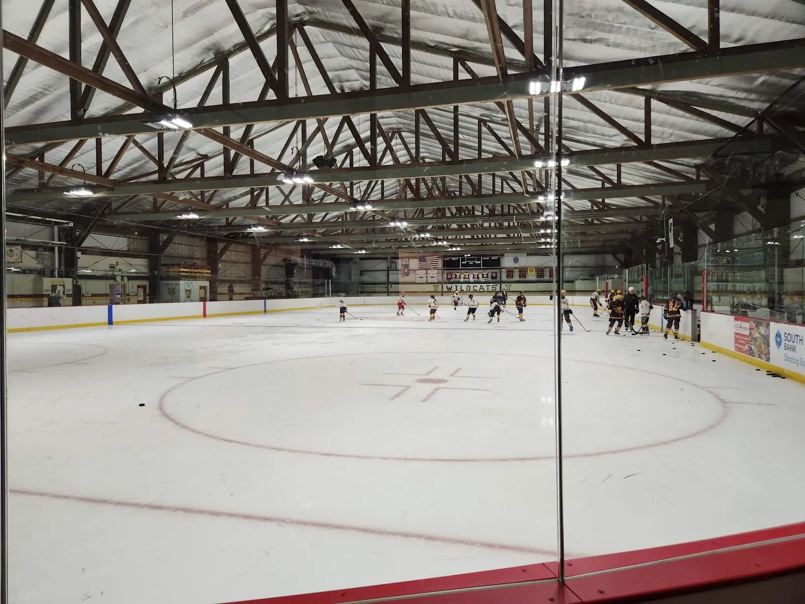 The ice sheet at the Connell rink under its timber and steel roof trusses, seen through the glass with a practice group far across the ice — Connell Memorial Rink