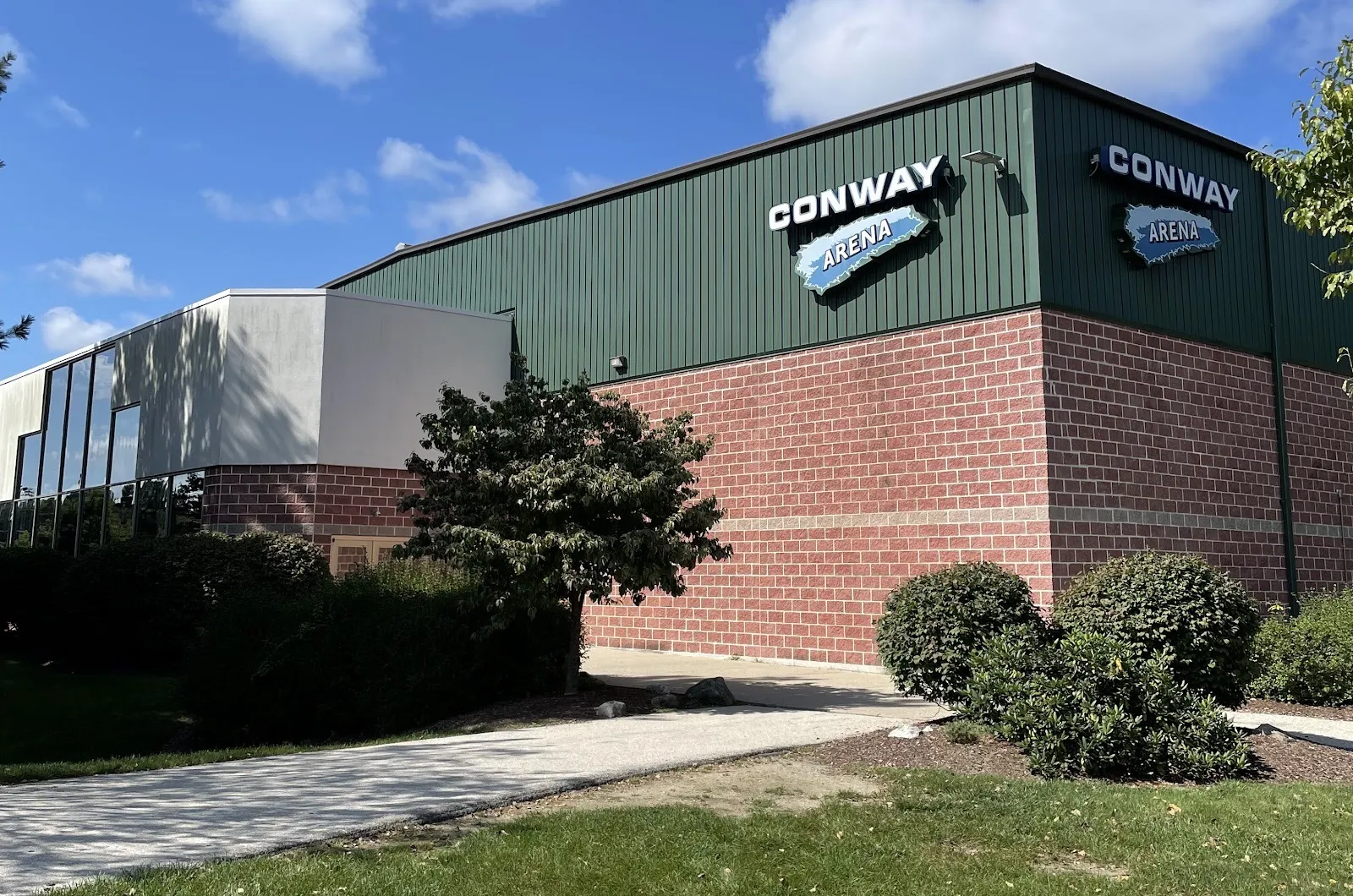 The green and red-brick Conway Arena on Stadium Drive in Nashua, with its name signs on two corners under a blue sky — Conway Arena
