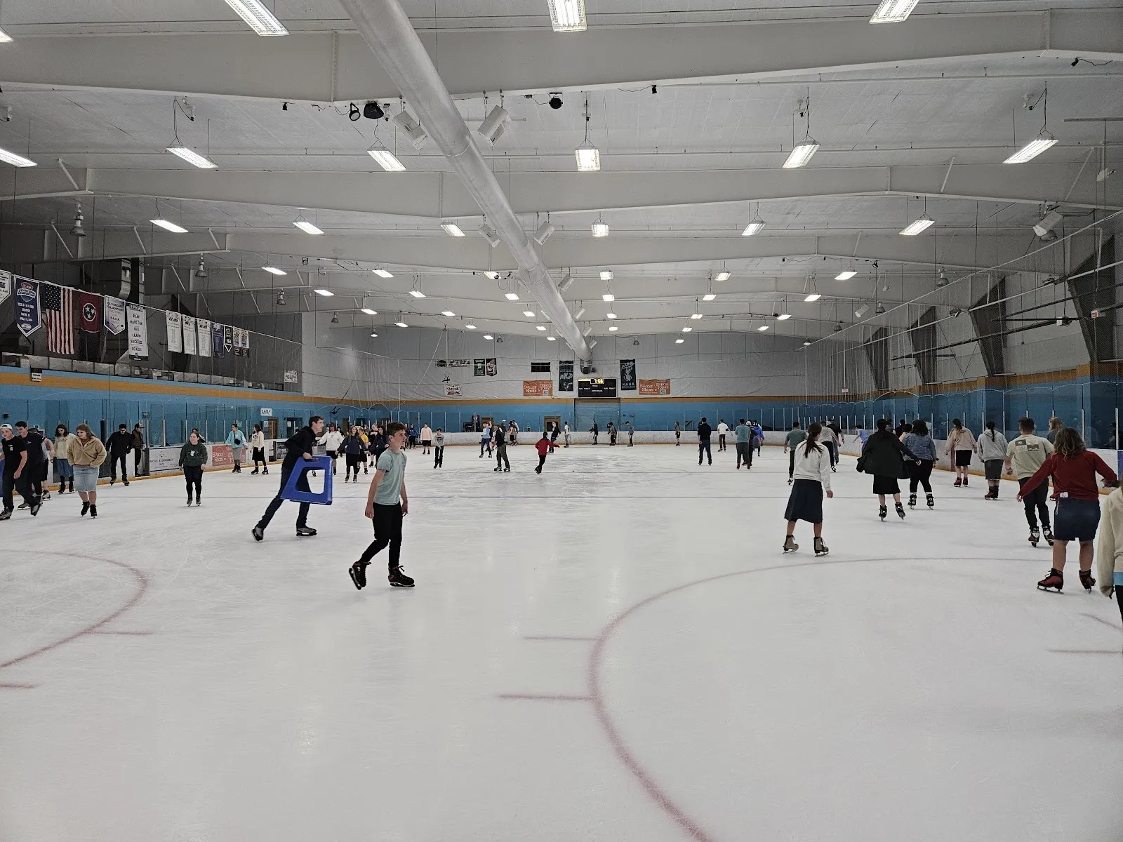 Una concurrida sesión pública en una pista amplia, con patinadores repartidos por el hielo bajo un alto techo blanco, con vallas azules y naranjas en la pared del fondo. — Cool Sports