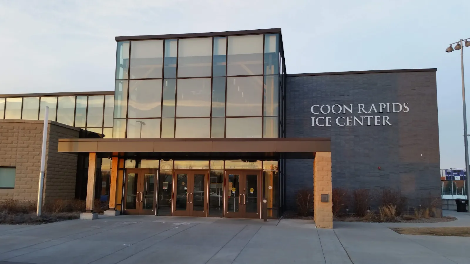 The glass-fronted entrance of the Coon Rapids Ice Center with its name on the dark wall, a timber canopy and a paved forecourt in evening light. — Coon Rapids Ice Center Outdoor Rink