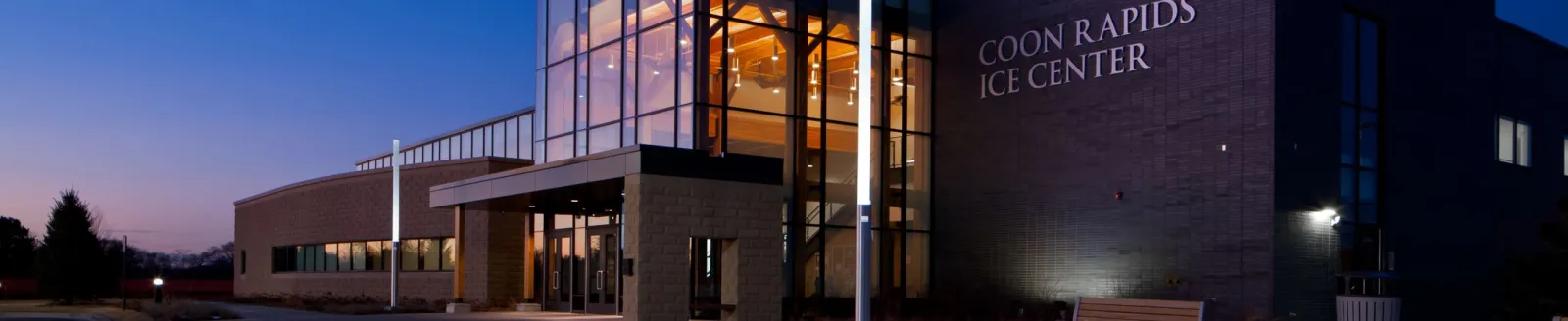 The building's glass-and-brick entrance at dusk, with illuminated 'COON RAPIDS ICE CENTER' wall lettering above the doors. — Coon Rapids Ice Center