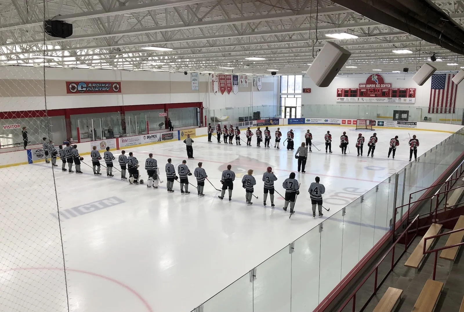 Two lines of players stand along the blue lines before a game, the wide sheet seen over the glass under a steel roof with a scoreboard and flags on the far wall. — Coon Rapids Ice Center