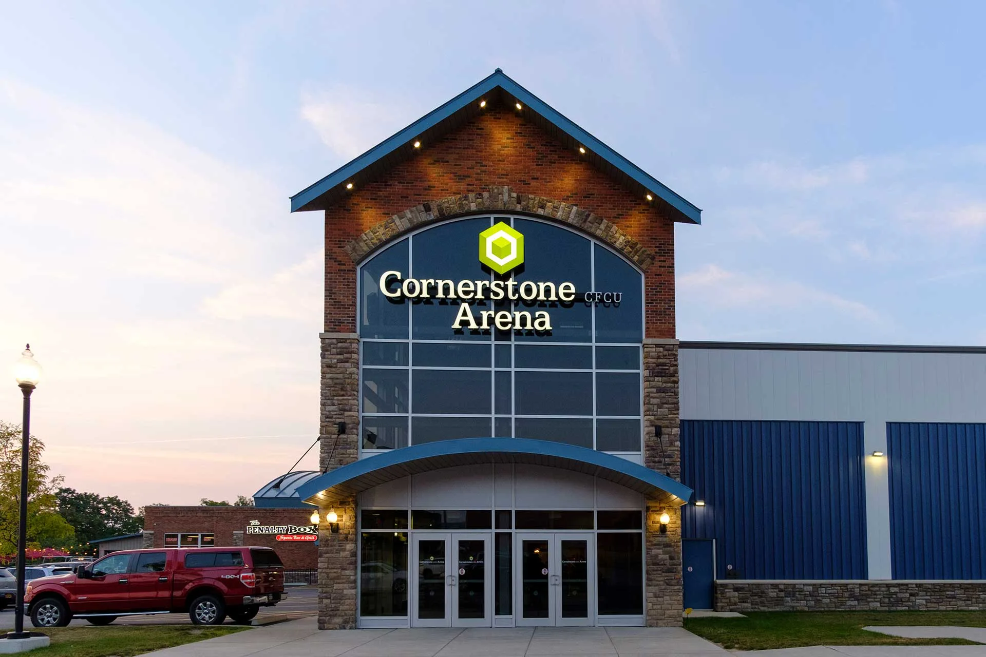 Frontón de la entrada al atardecer, iluminado desde abajo bajo un techo inclinado con bordes azules — Cornerstone CFCU Arena