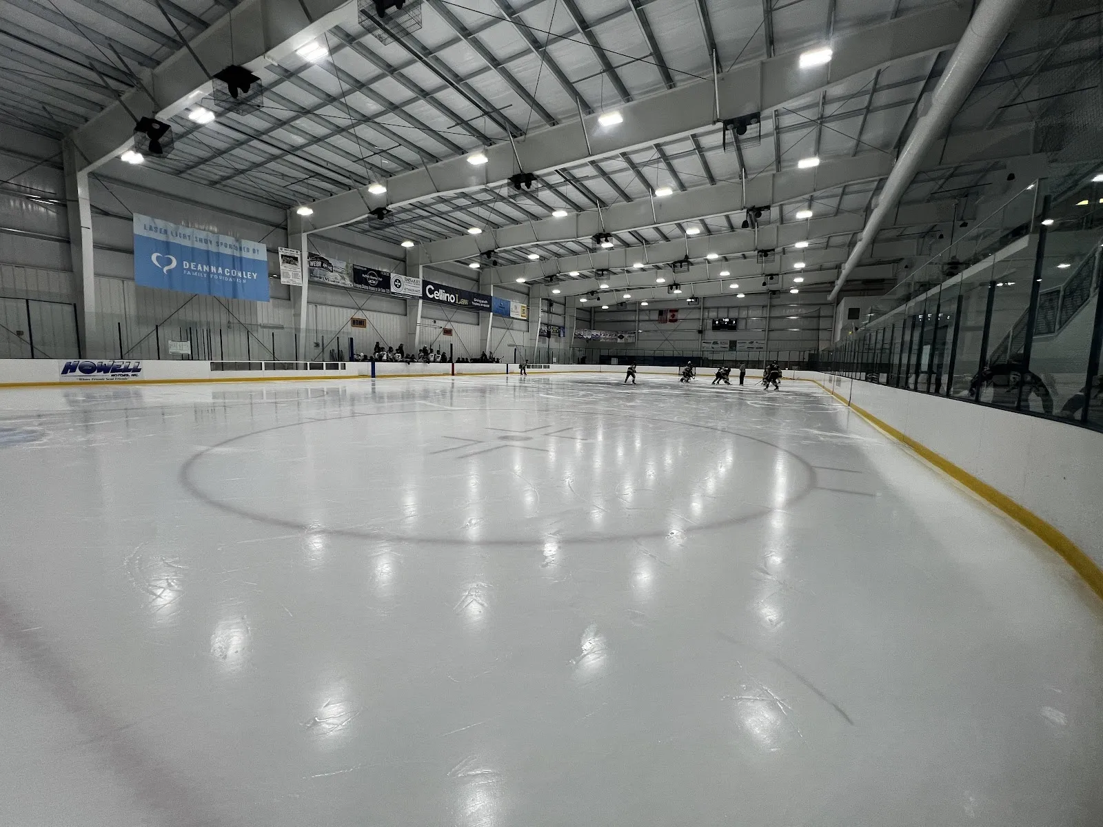 Una pista vacía vista desde la esquina de las vallas, con hielo blanco, un círculo central y vallas de borde amarillo bajo un alto techo de armaduras de acero. — Cornerstone CFCU Arena