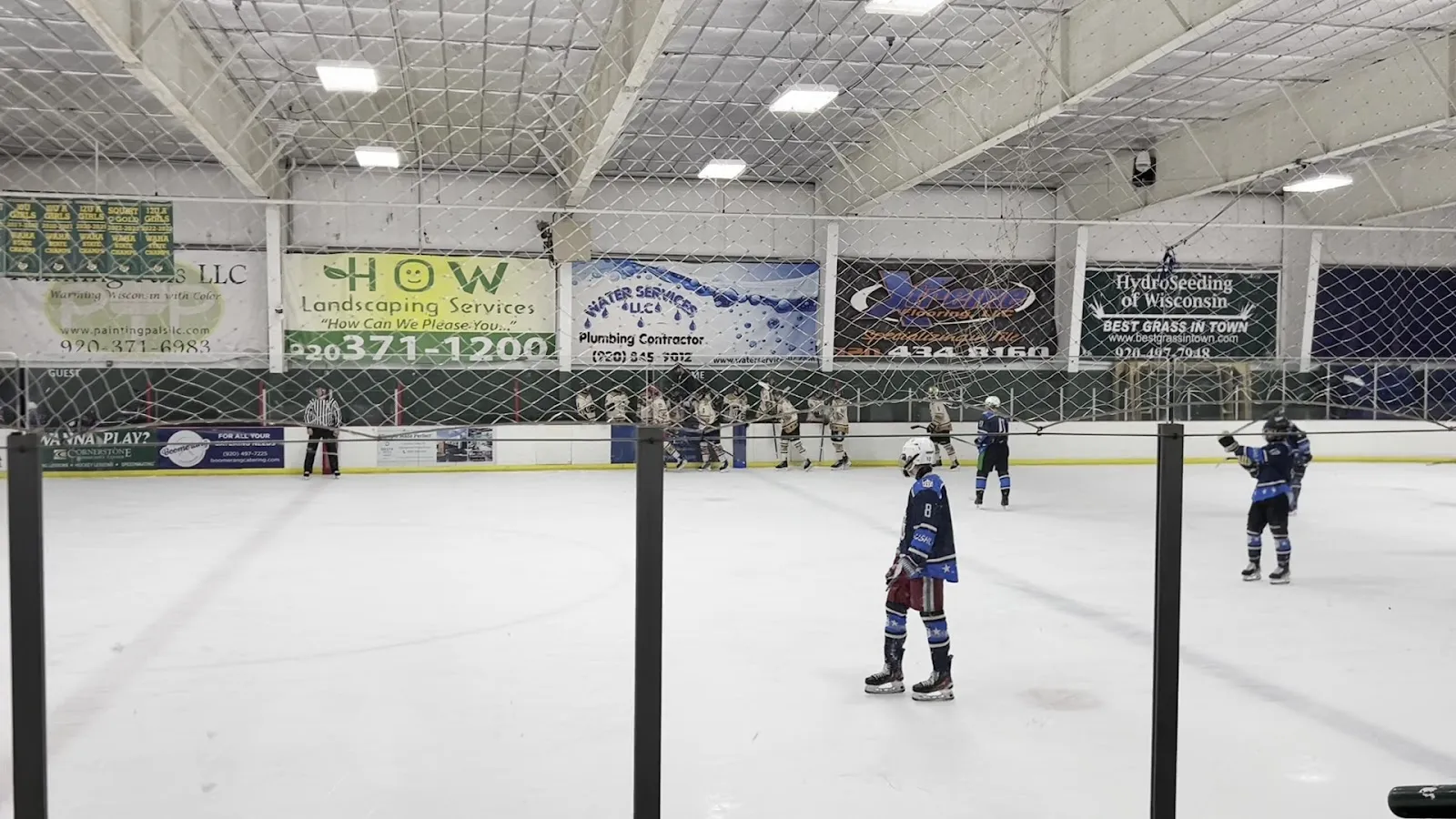 A wide view of the ice from the glass, a few skaters spread along the far side beneath a high ceiling with sponsor boards and a long scoreboard on the end wall. — Cornerstone Community Center