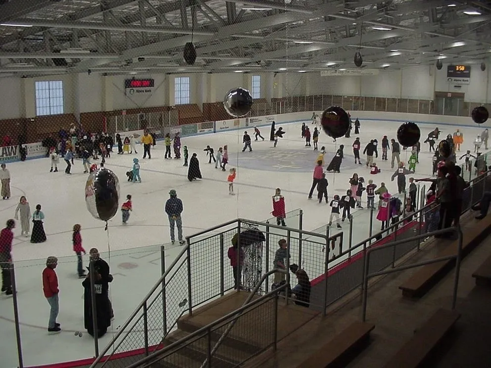 A public skating session across a wide sheet, children and families skating and gathering among black balloons beneath a steel-trussed roof with a scoreboard on the far wall. — Cornerstone Community Center