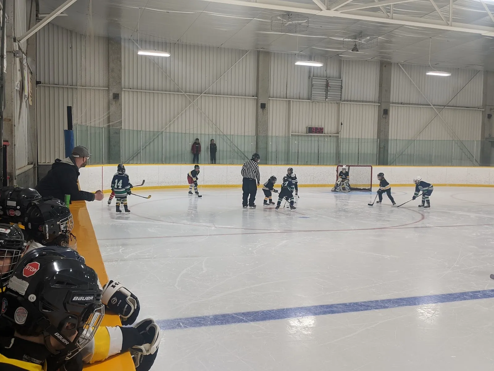 A practice at one end of the sheet seen from the glass, players spread across the ice under a long steel-framed grey roof. — Coronation Arena (Hamilton)