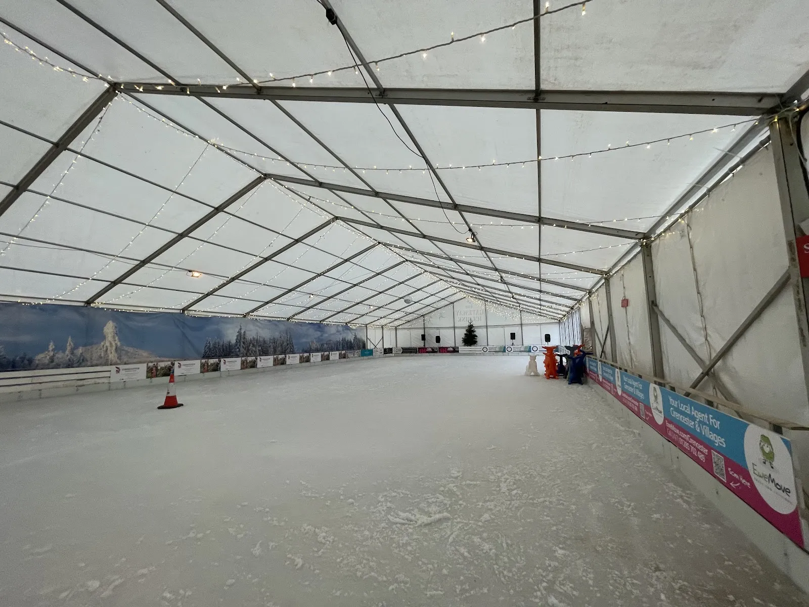 The empty ice sheet inside the long white marquee of the Cotswold Ice Rink at the Cotswold Water Park, with string lights and a painted alpine backdrop along the boards — Cotswold Ice Rink