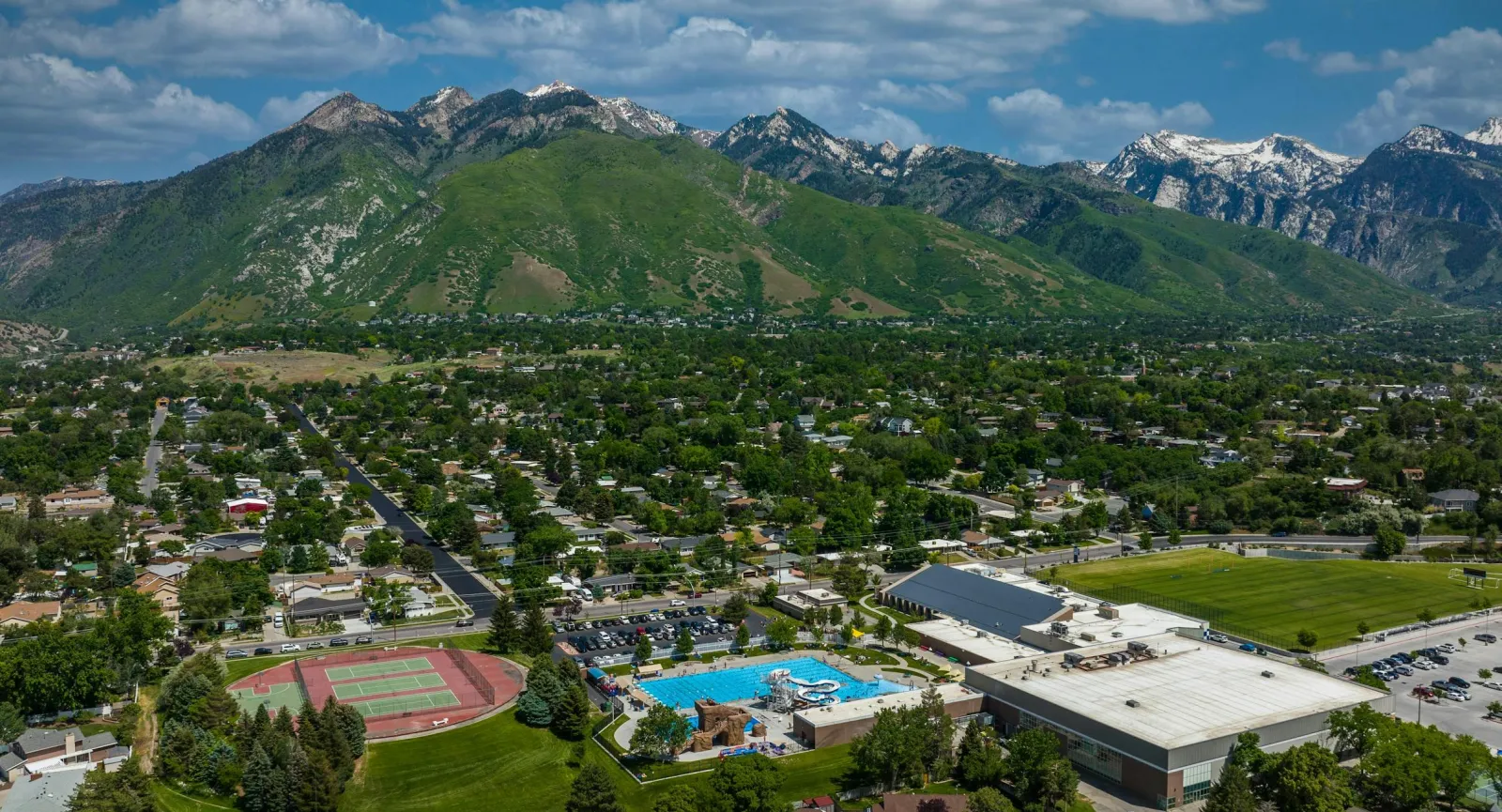 El centro recreativo visto desde el aire, con su largo edificio de techo plano junto a las albercas exteriores y las canchas de tenis, y las montañas Wasatch al fondo. — Cottonwood Heights Ice Arena